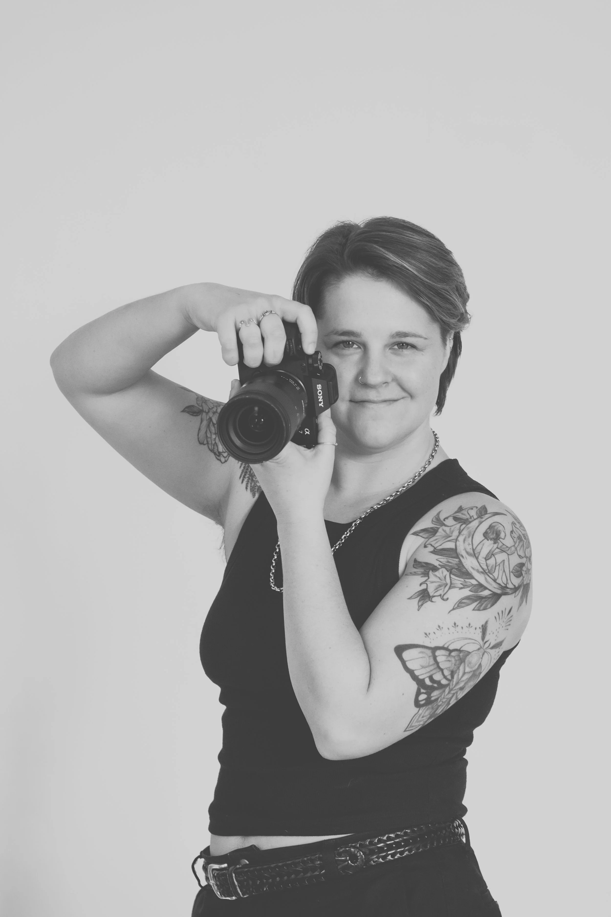 black and white image of queer photographer holding camera in front of their face. arms have tattoos.