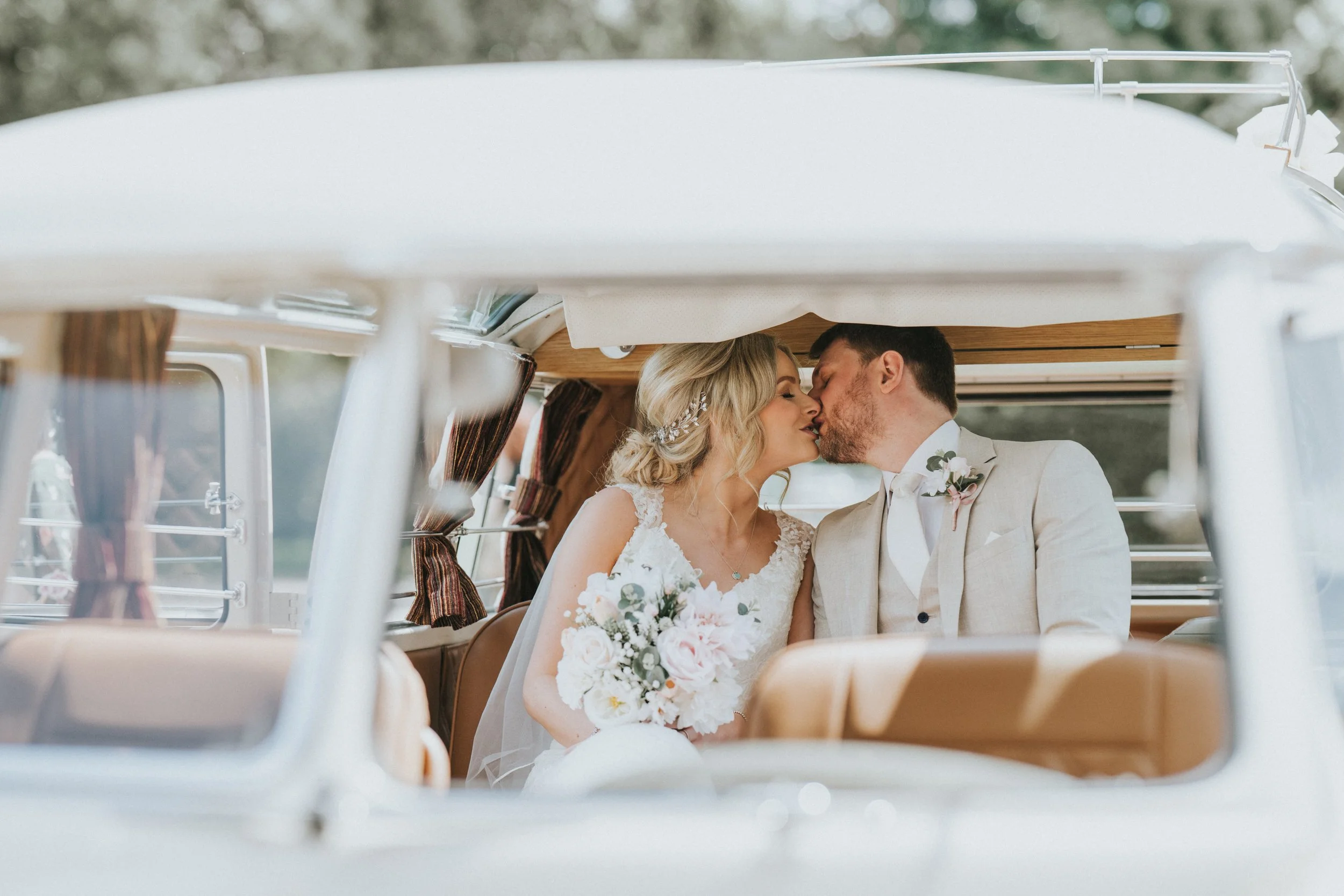 A bride and groom sharing a kiss during their wedding ride in a vintage vehicle, with the bride holding a bouquet of flowers.
