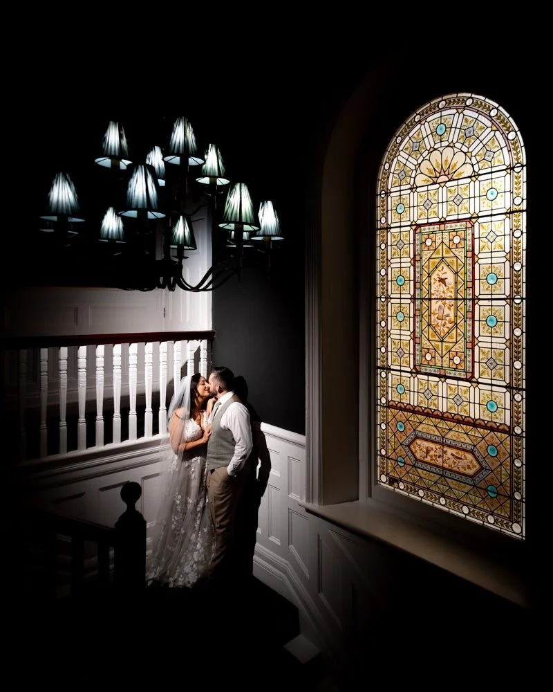 A bride and groom sharing a kiss on a staircase inside a building with a large, ornate stained-glass window to their right and a chandelier hanging above them.