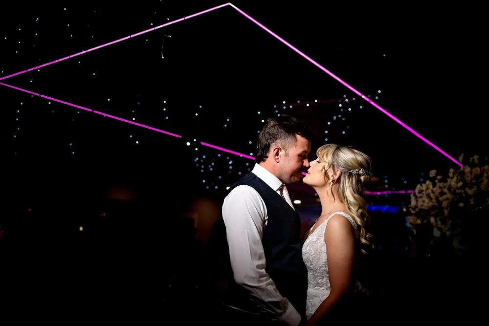 A couple dressed in wedding attire standing close together, about to kiss, with a dark background illuminated by pink and purple neon lights and small decorative lights.
