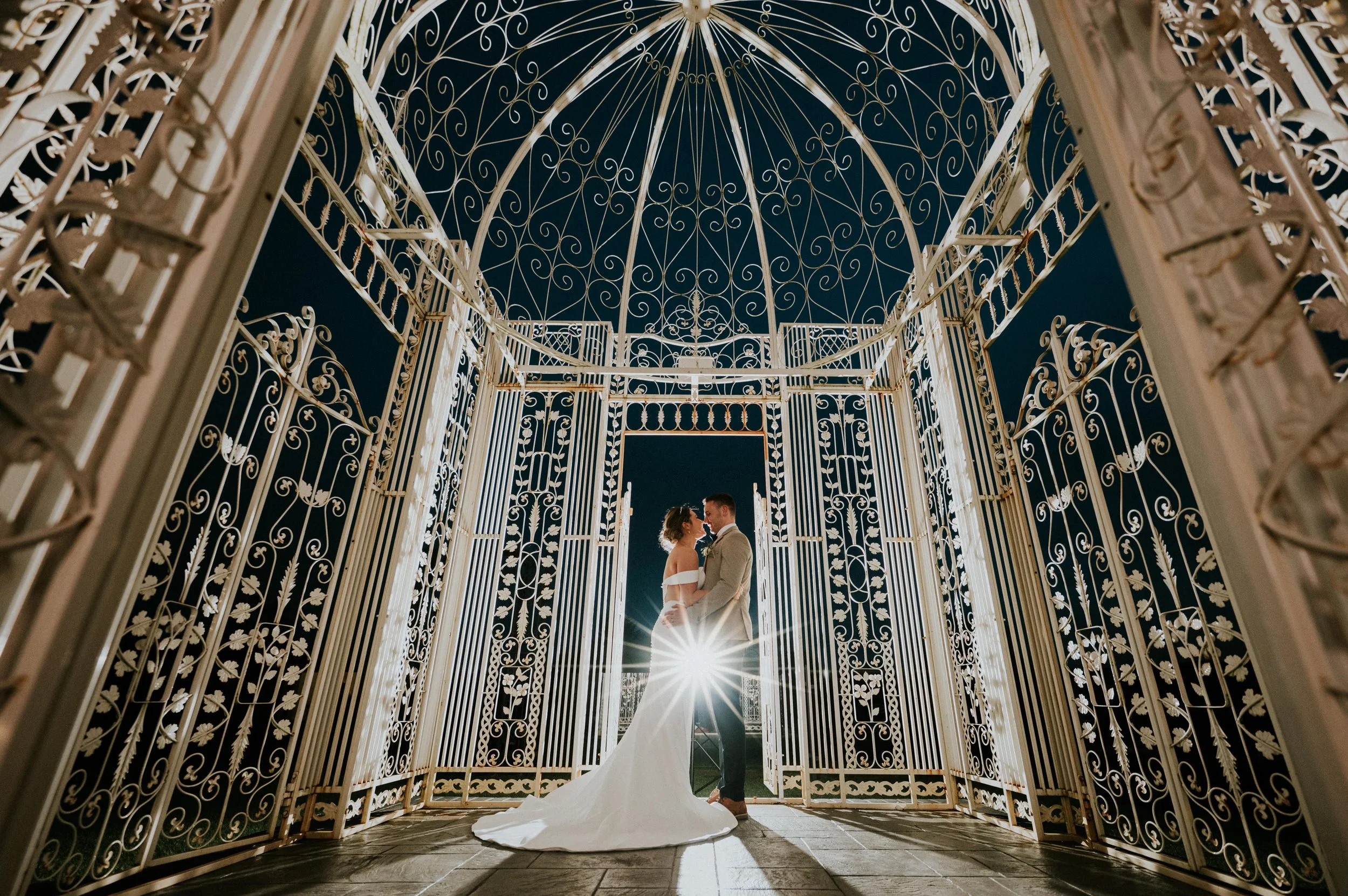 A bride and groom standing close together in a decorative pavilion at night, with sunlight shining from behind, creating a dramatic silhouette effect.