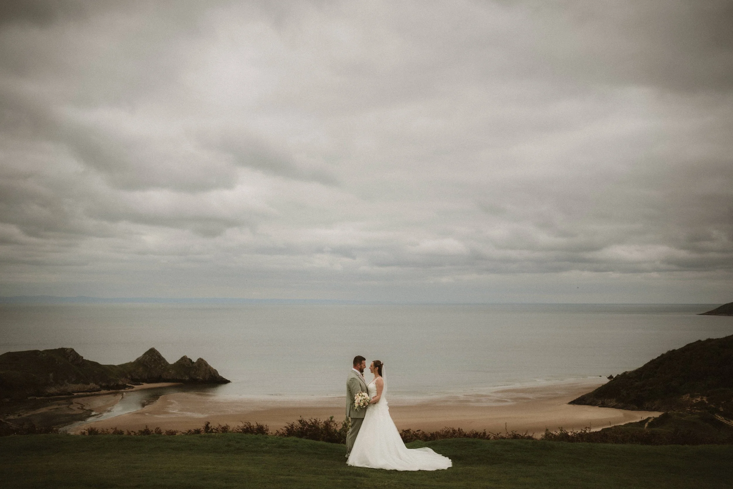 A bride and groom standing on a grassy area near the beach on a cloudy day, facing each other and holding a bouquet of flowers, with ocean and cliffs in the background.