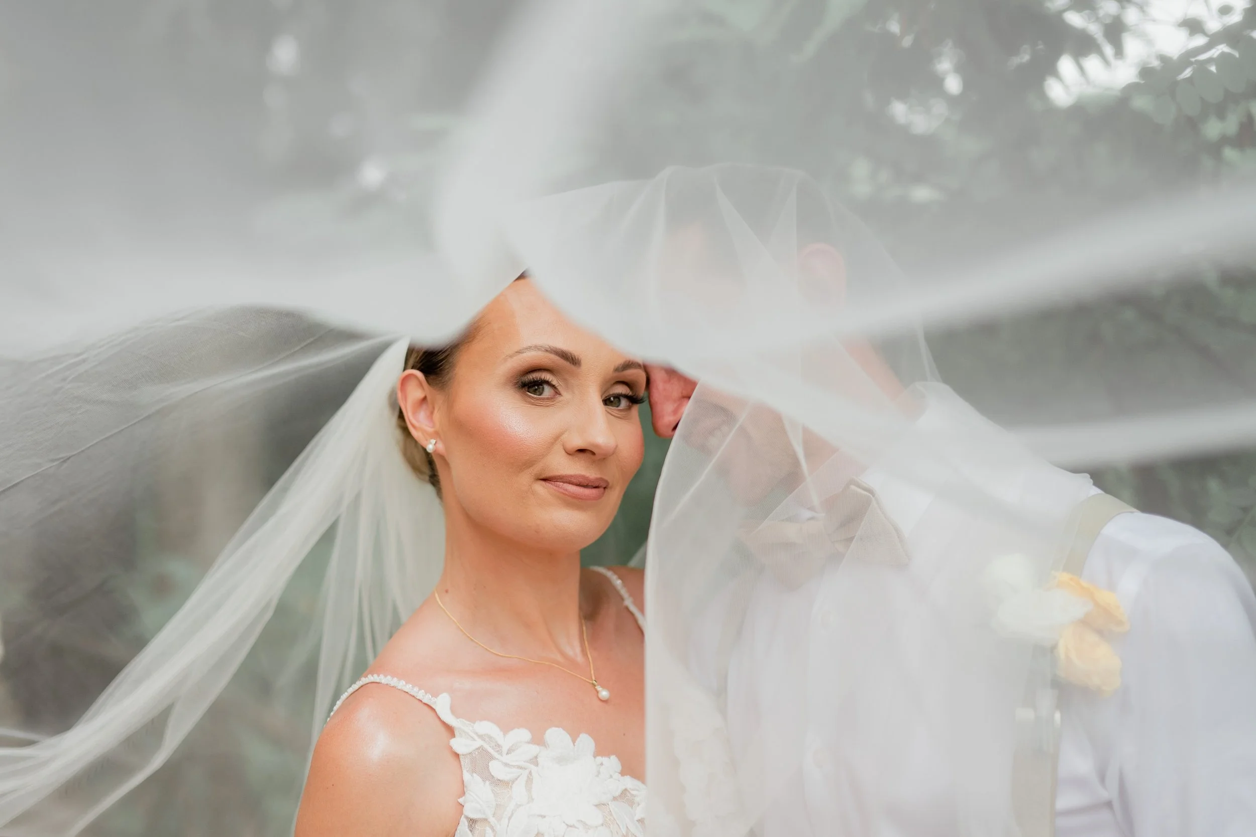 A bride with makeup and earrings, wearing a lace wedding dress and veil, smiling behind sheer fabric.