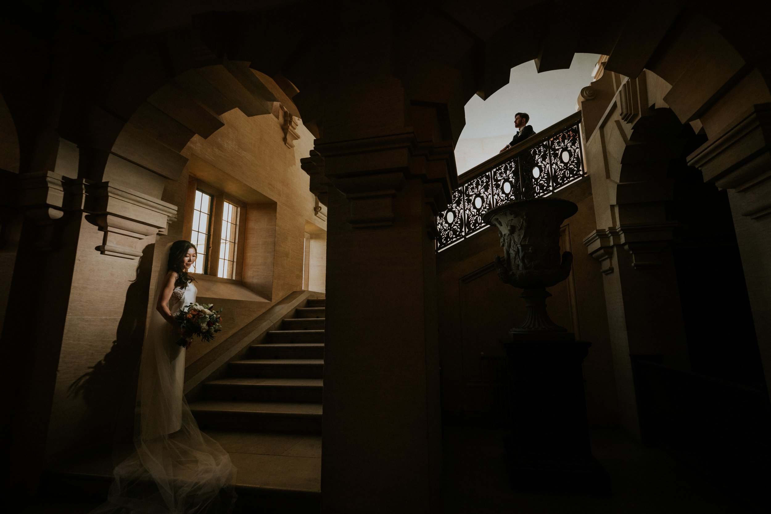 A bride holding a bouquet of flowers stands on a staircase inside a grand building, with a groom standing on the balcony above, gazing down at her.
