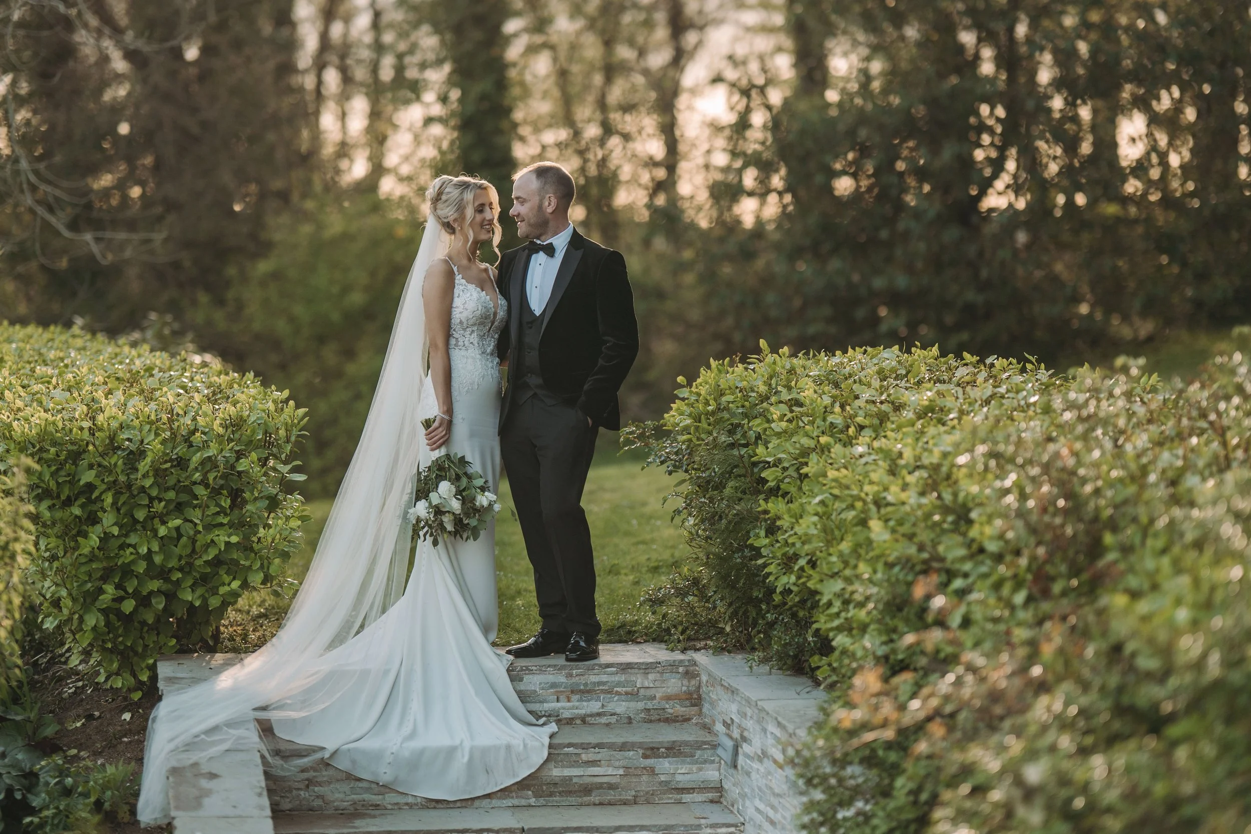A bride and groom standing closely together outdoors during sunset, smiling and looking at each other. The bride is wearing a white wedding dress with a long train and veil, holding a bouquet. The groom is dressed in a black tuxedo with a bow tie. Th