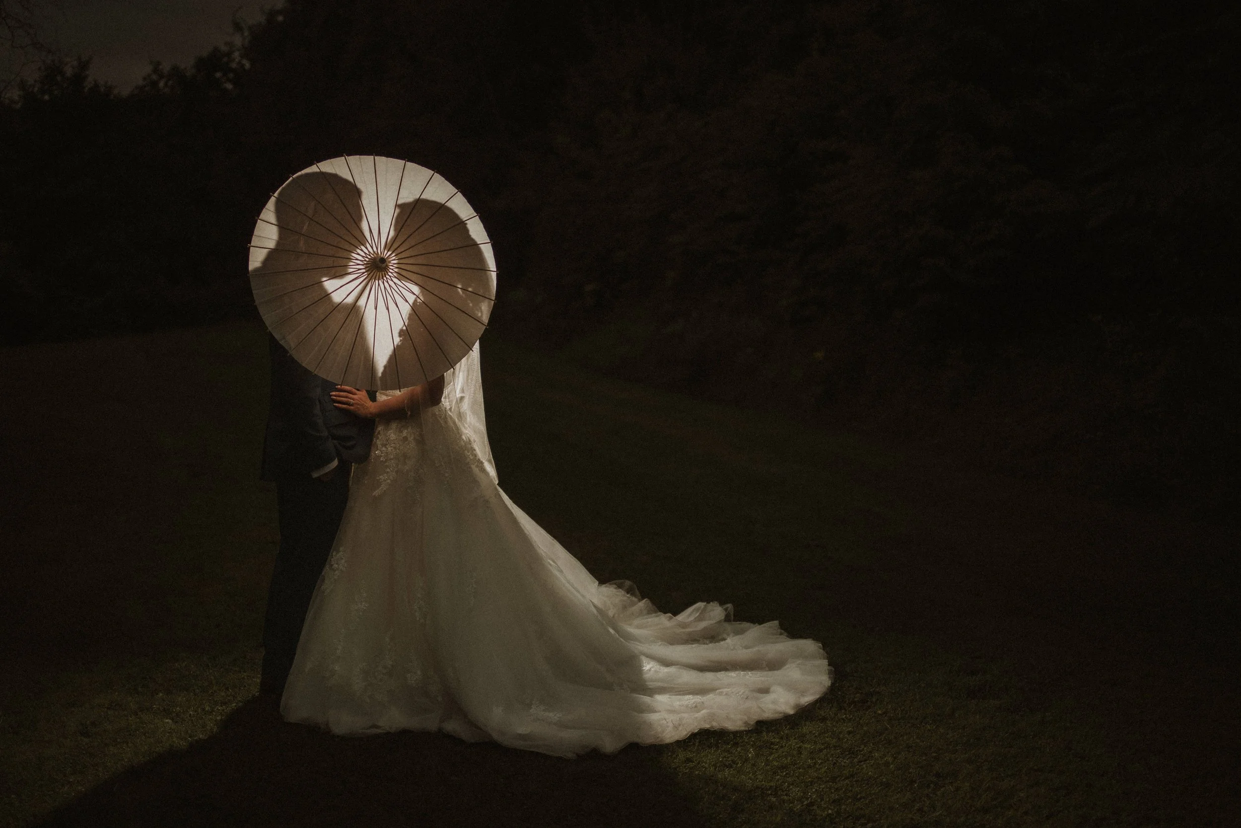 A couple, possibly newlyweds, holding a white umbrella at night in an outdoor setting, with the woman wearing a wedding dress and the man in dark clothing, sharing an intimate moment.