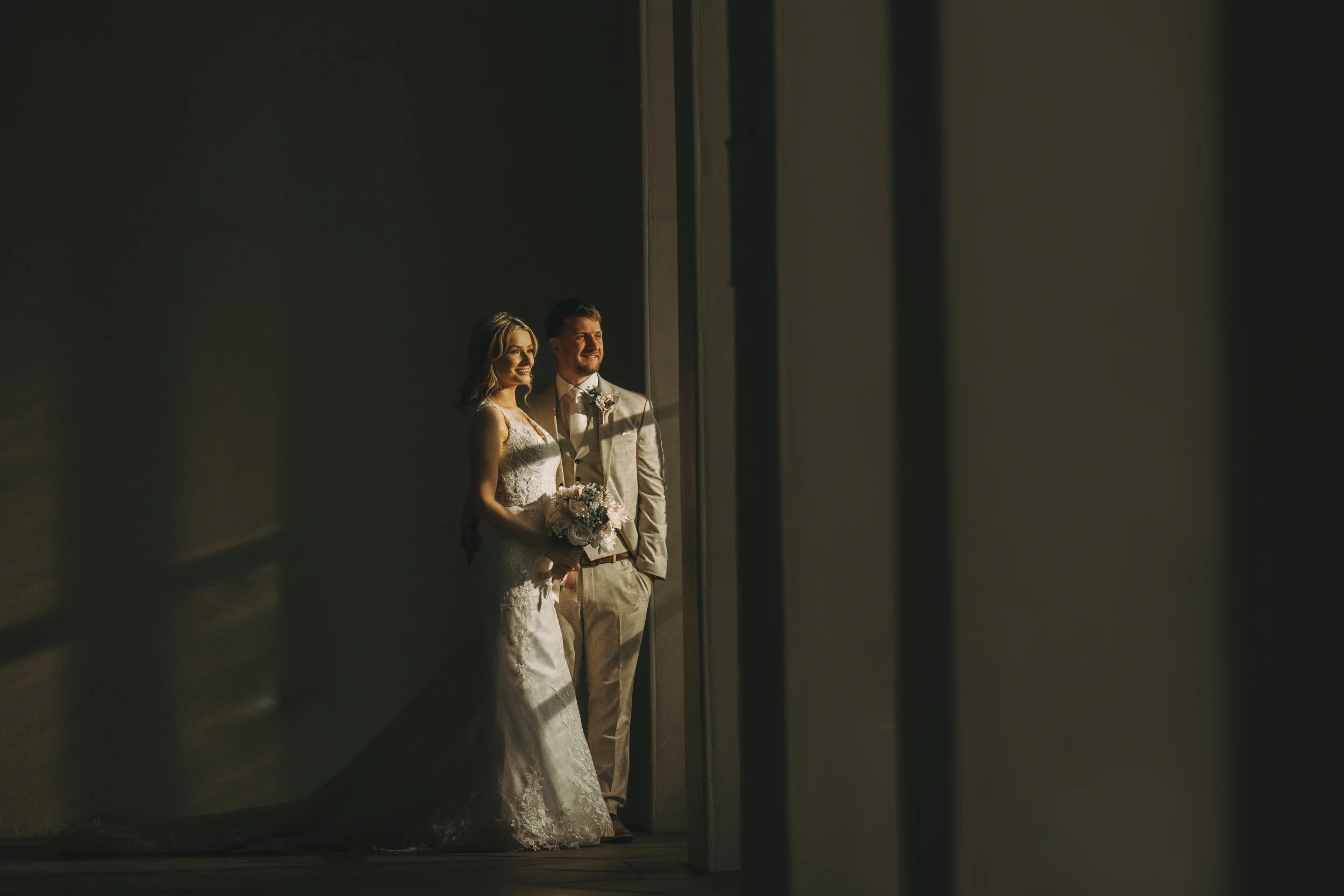 A bride and groom standing by large windows, dressed in wedding attire, illuminated by soft natural light.