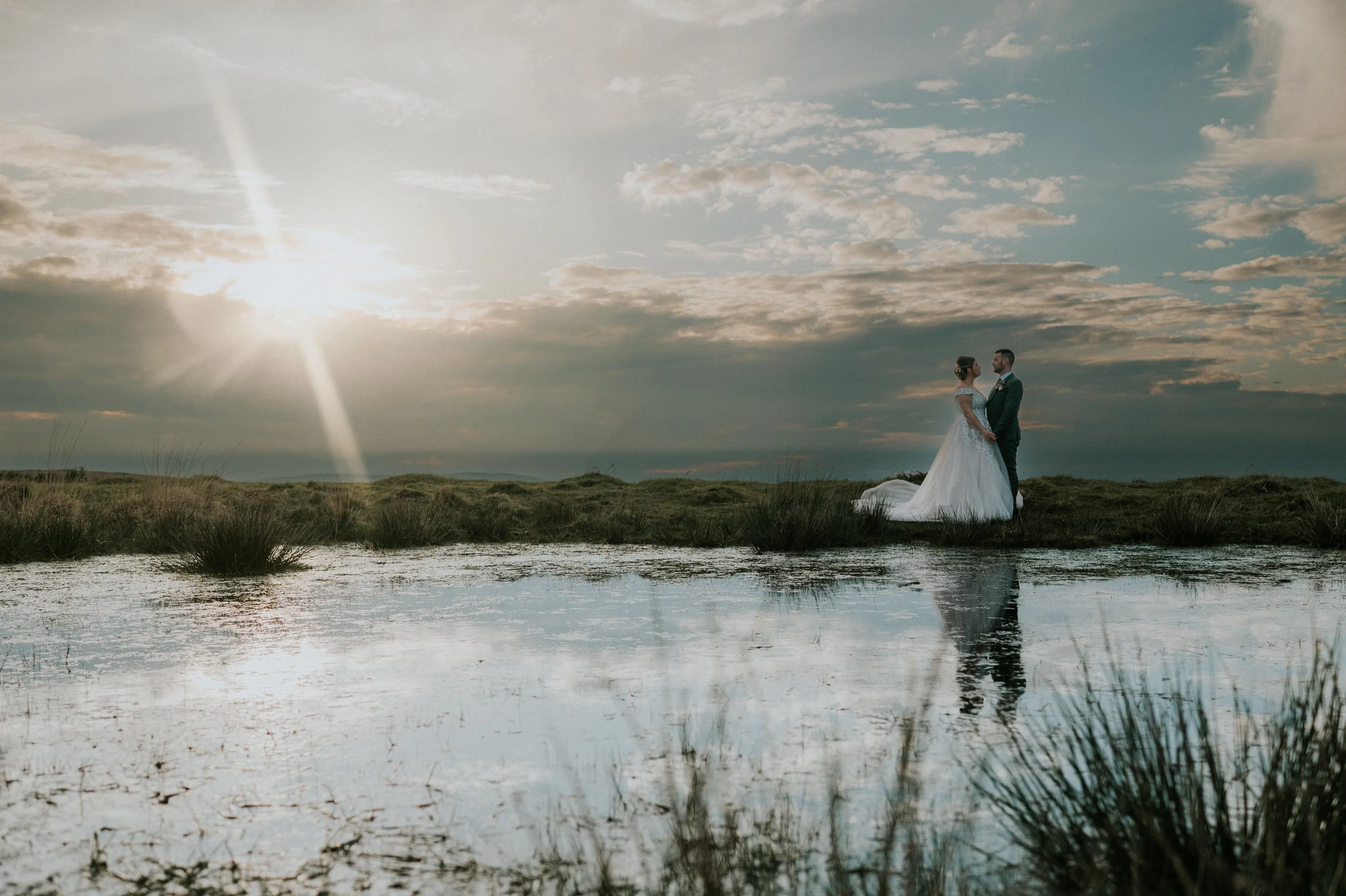 A bride and groom standing face to face on a grassy landscape with a pond, under a cloudy sky during sunset.