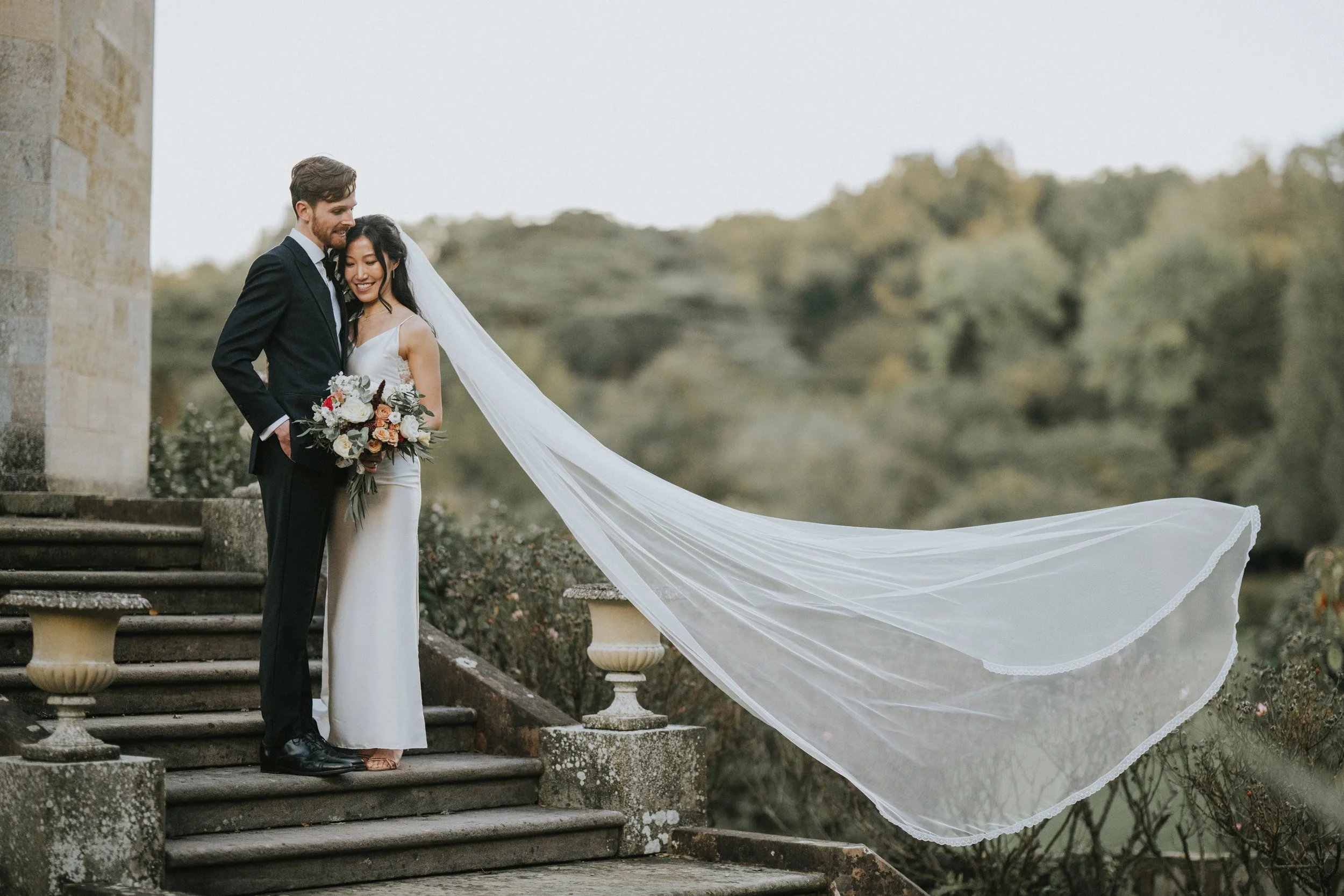A bride and groom standing on stone steps outdoors, with the bride holding a bouquet and both smiling, the bride's veil flowing in the breeze.