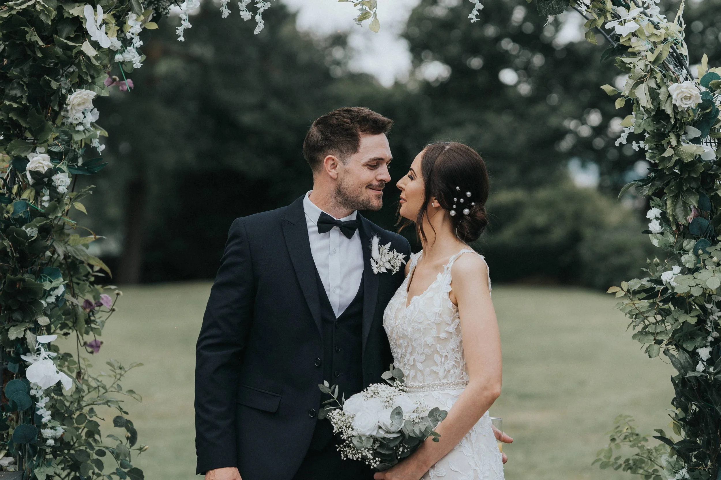 A bride and groom standing close together during their outdoor wedding ceremony, framed by greenery and flowers, gazing at each other lovingly.