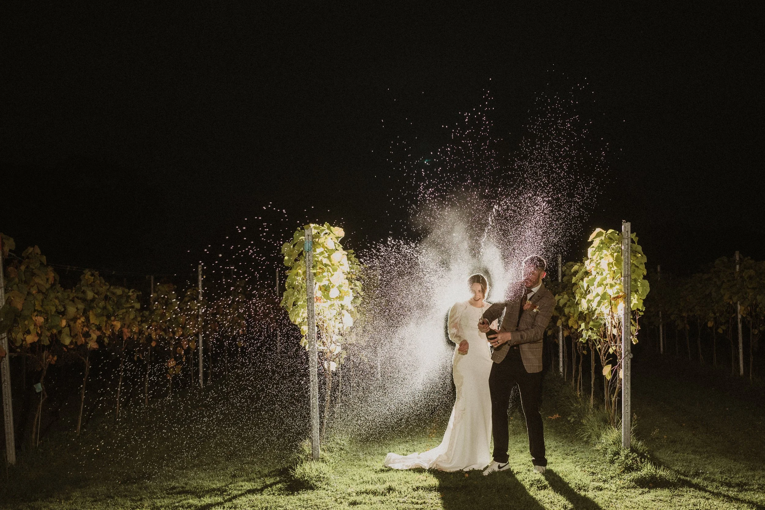 A bride and groom celebrating at night in a vineyard, standing under sparklers with grapevines on either side.
