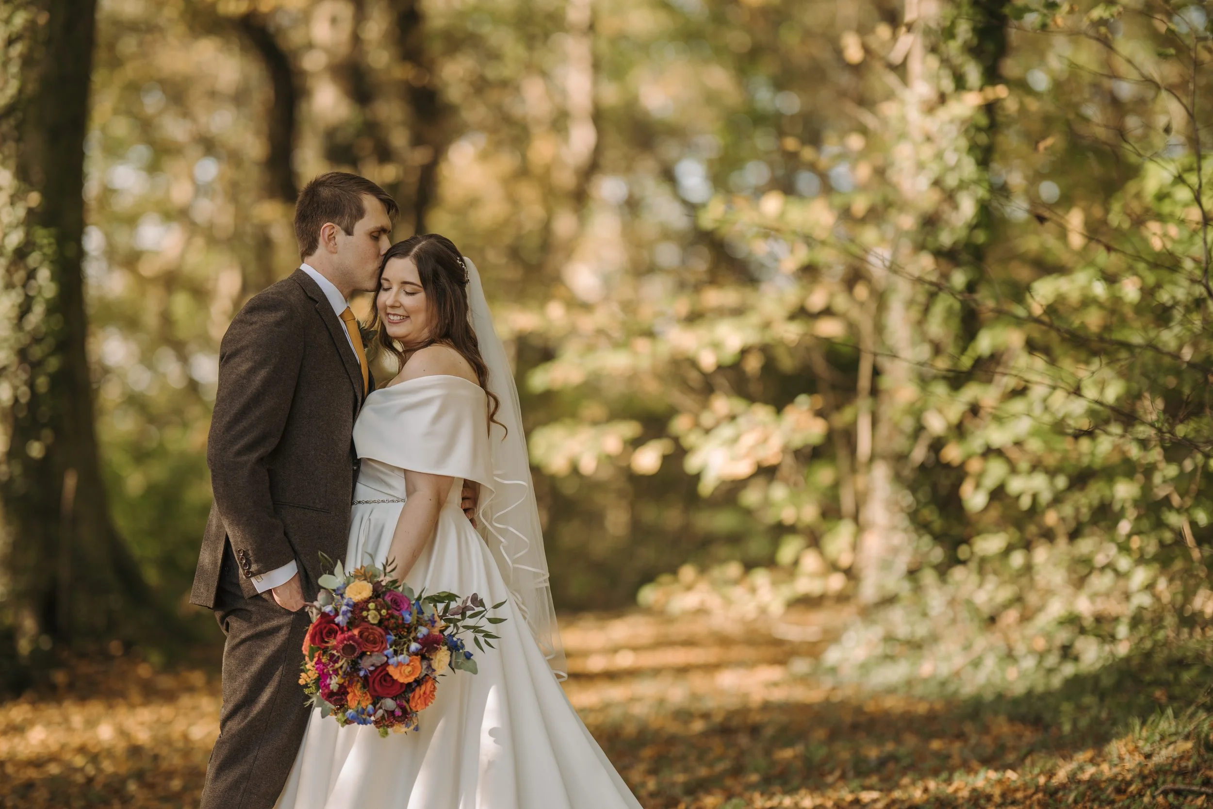 A bride and groom in a forest setting, the groom is kissing the bride on the forehead, both are smiling, the bride holds a colorful bouquet, and autumn leaves are visible on the ground.