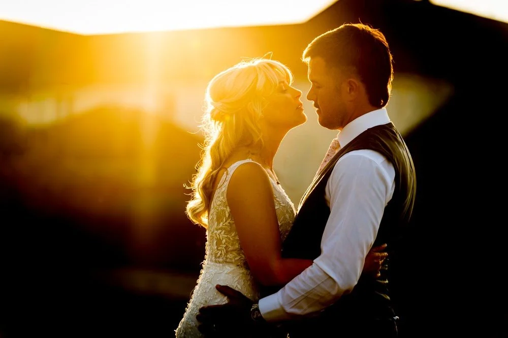 A couple in wedding attire sharing an intimate moment at sunset, facing each other with faces close, with a warm glow in the background.