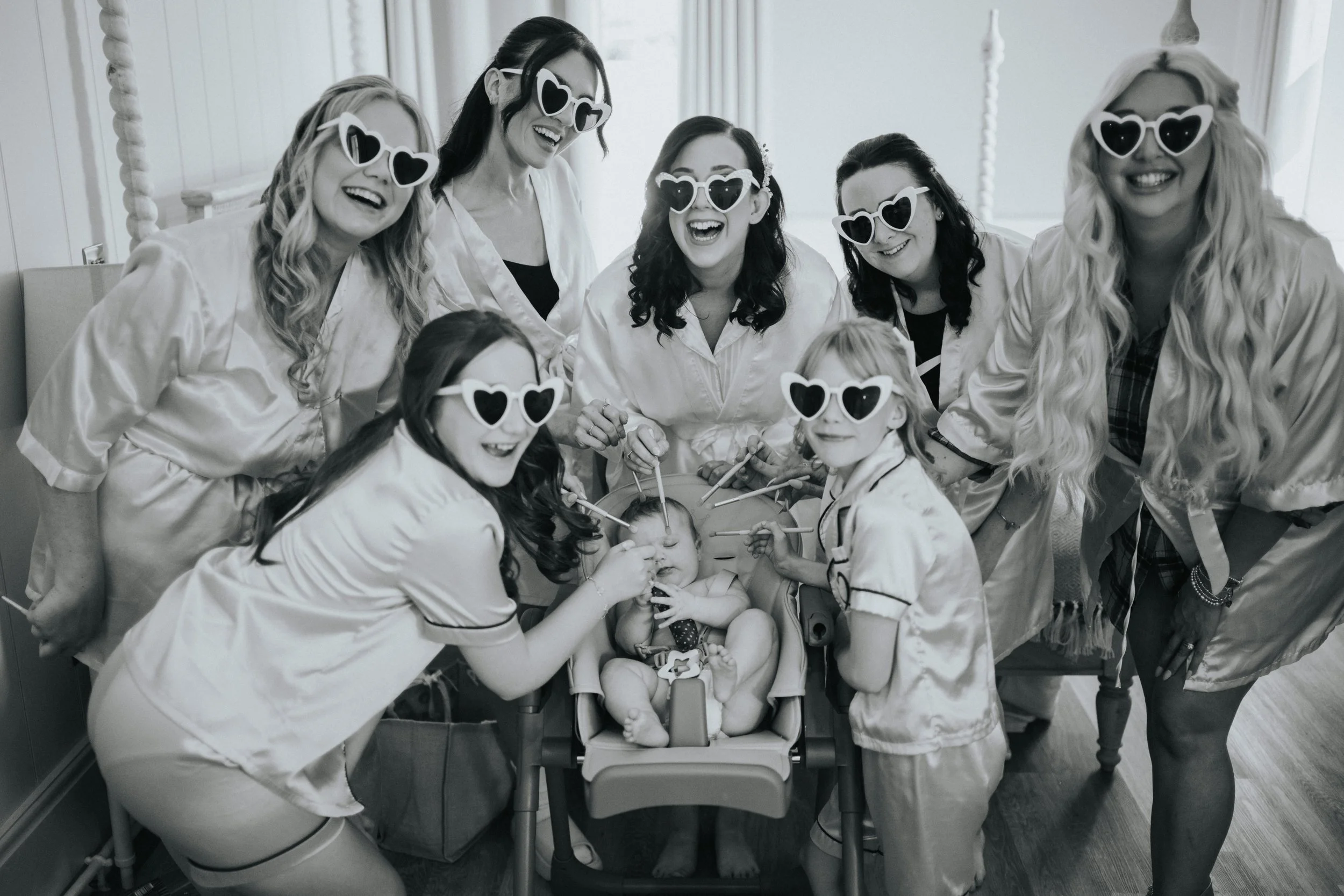 Group of women wearing white robes and heart-shaped sunglasses gathered around a baby in a high chair, playing with the baby with sticks or tools, all smiling and having fun.