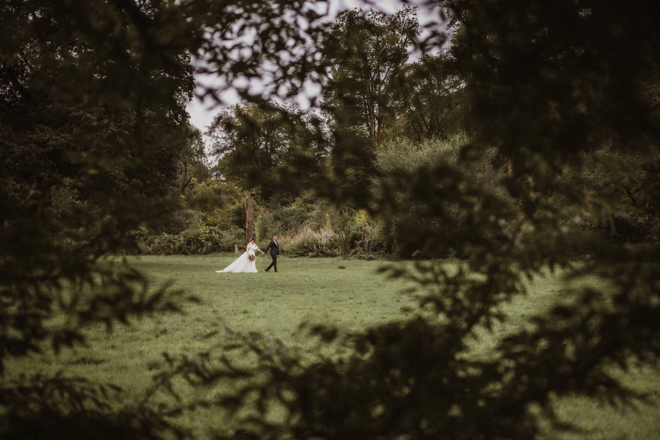 A bride and groom walking hand in hand on a grassy field, framed by trees and dense foliage.