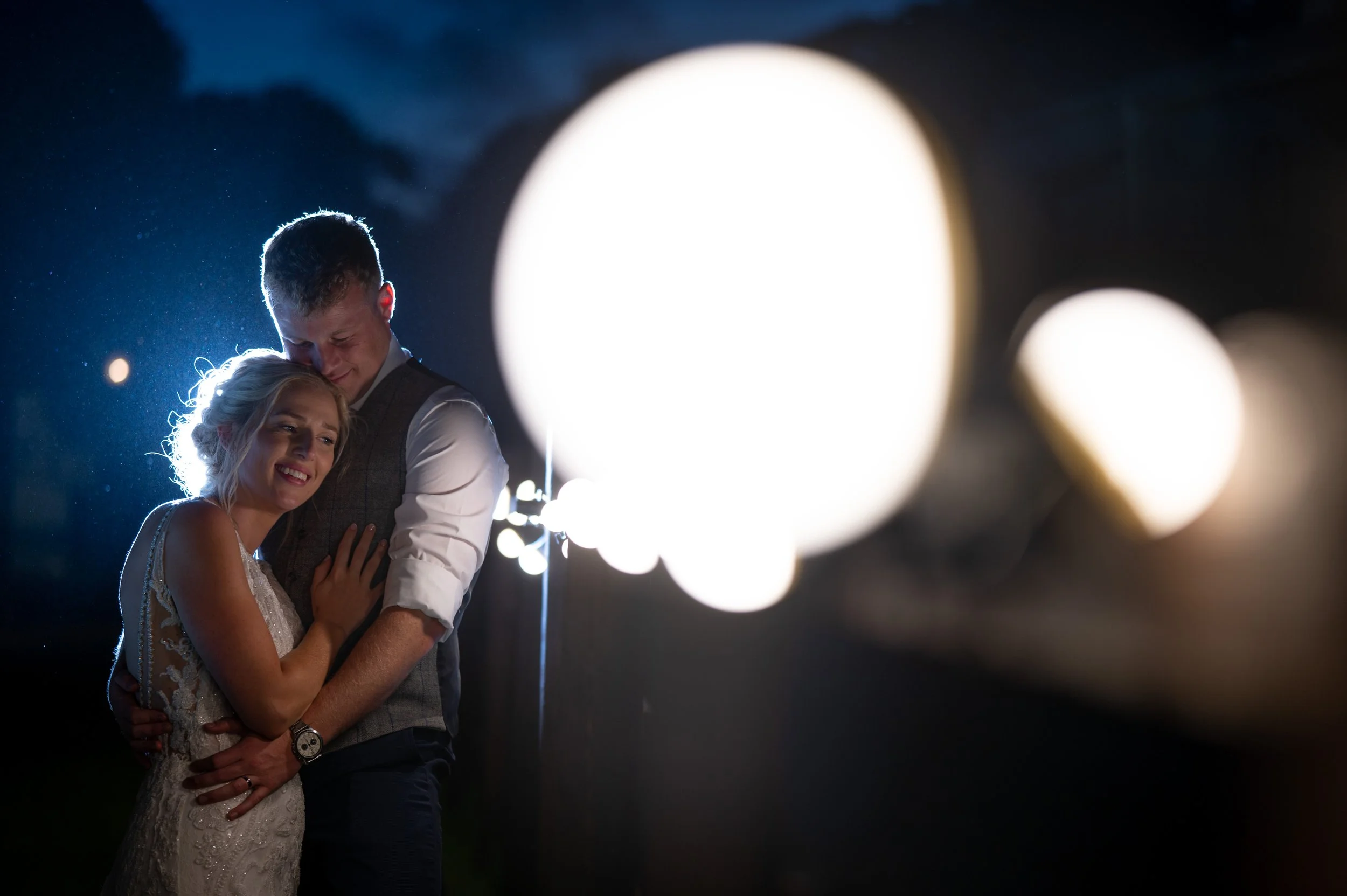 A couple embracing during a nighttime event, with bright lights blurred in the background.