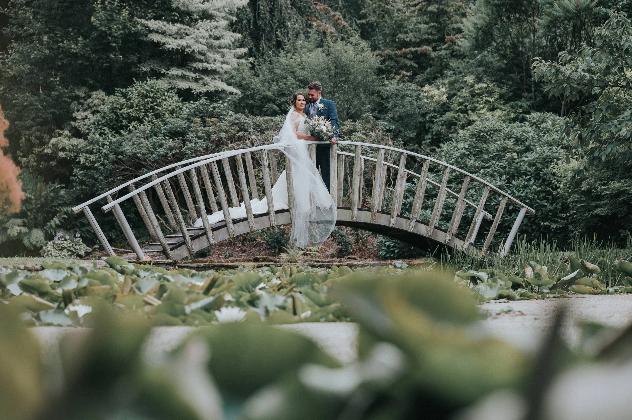 A bride and groom standing together on a small wooden bridge surrounded by lush greenery, with the bride holding a bouquet of flowers.