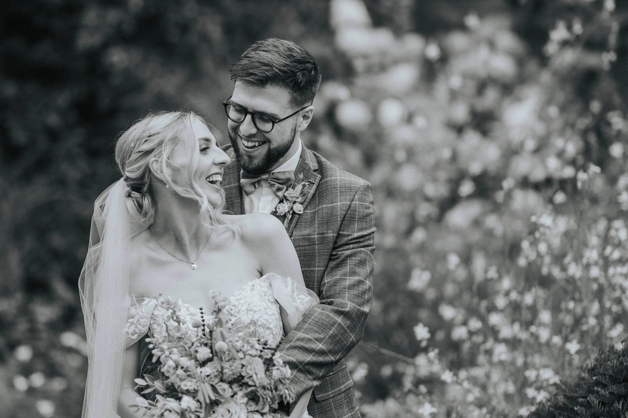 Black and white photo of a newlywed couple outdoors. The bride is holding a large bouquet and wearing a wedding dress, and the groom, in a checkered suit and glasses, is smiling and embracing her.