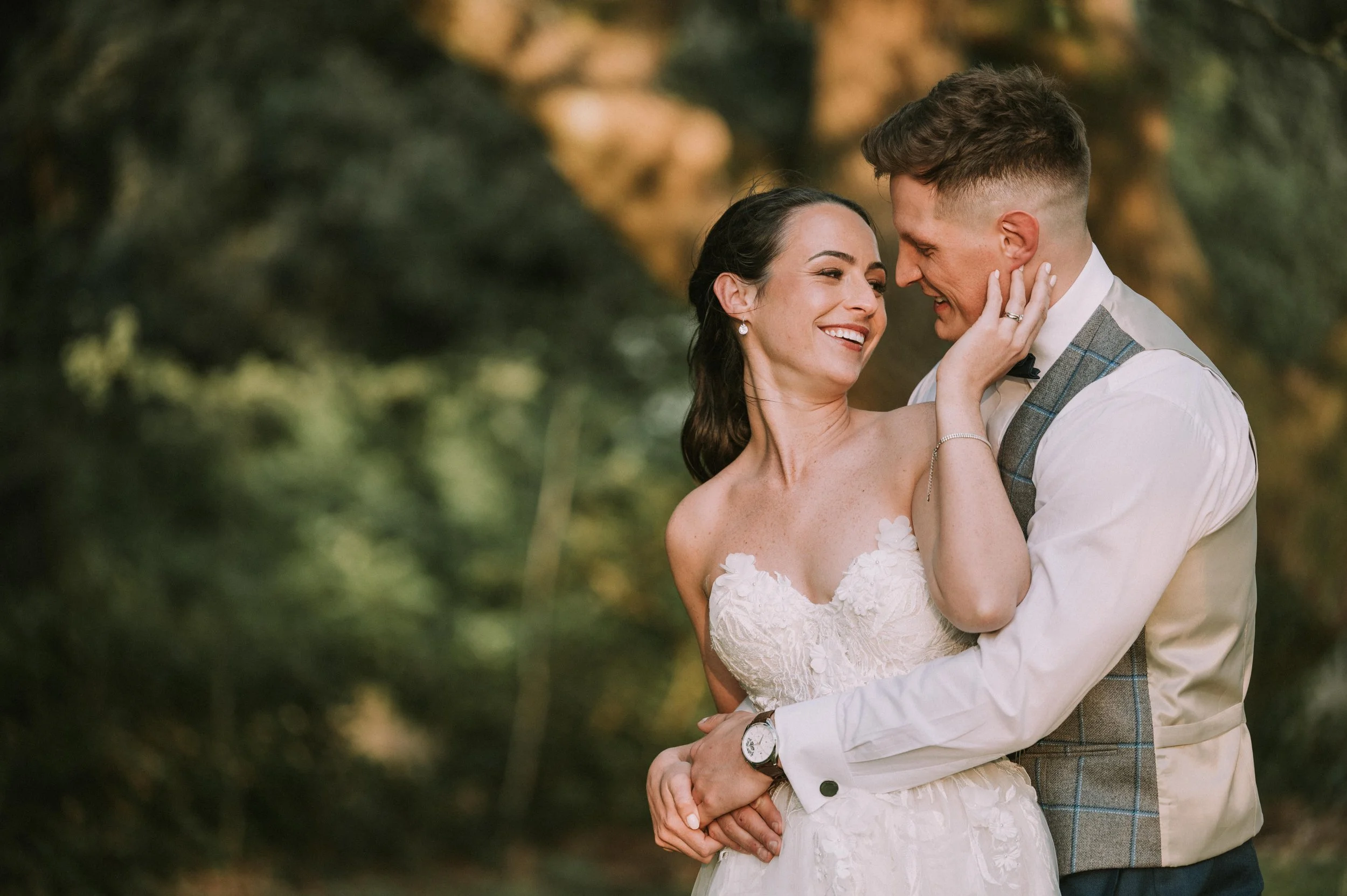 A newlywed couple sharing a joyful moment outdoors, with the woman in a white wedding dress and the man in a formal vest and shirt, surrounded by blurred greenery.