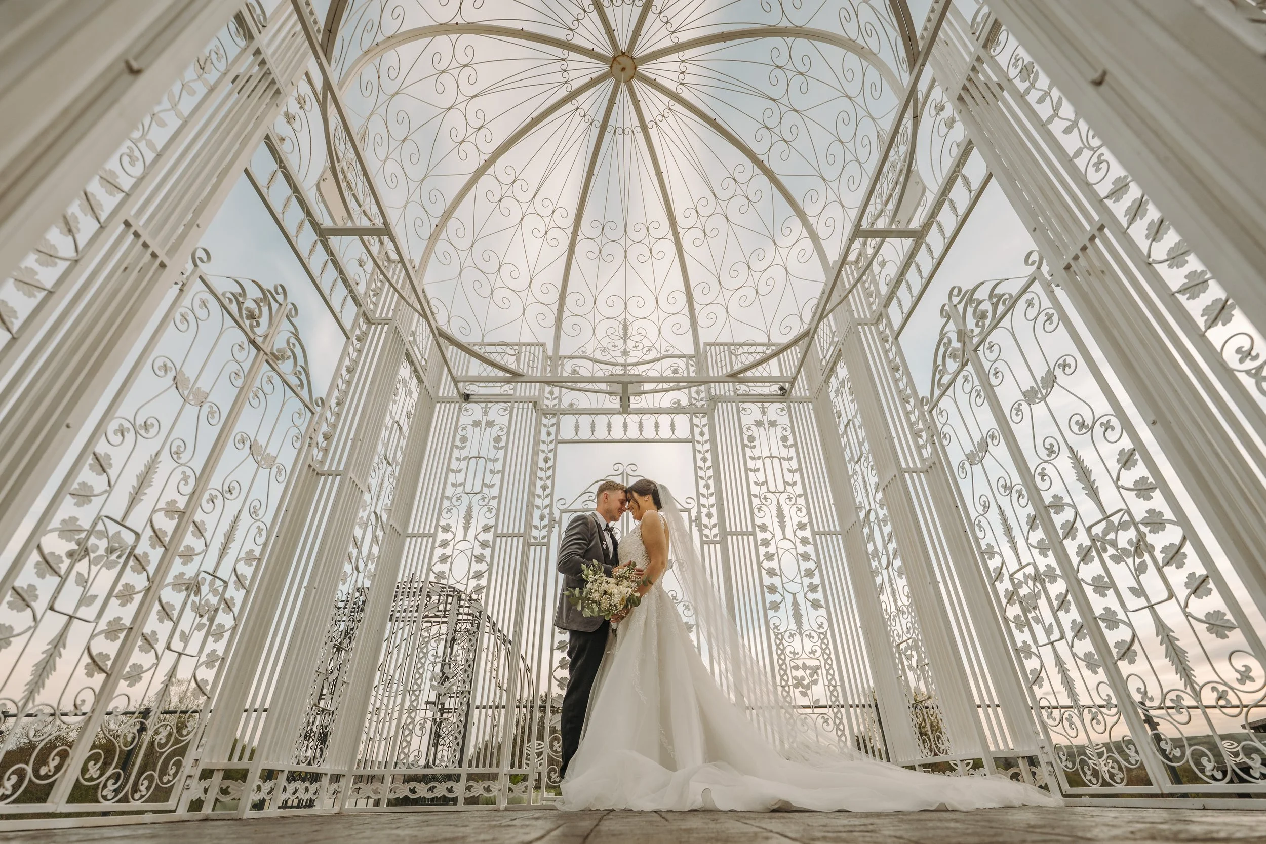 A bride and groom standing close together inside a decorative white gazebo, touching foreheads and holding a bouquet, on a cloudy day.