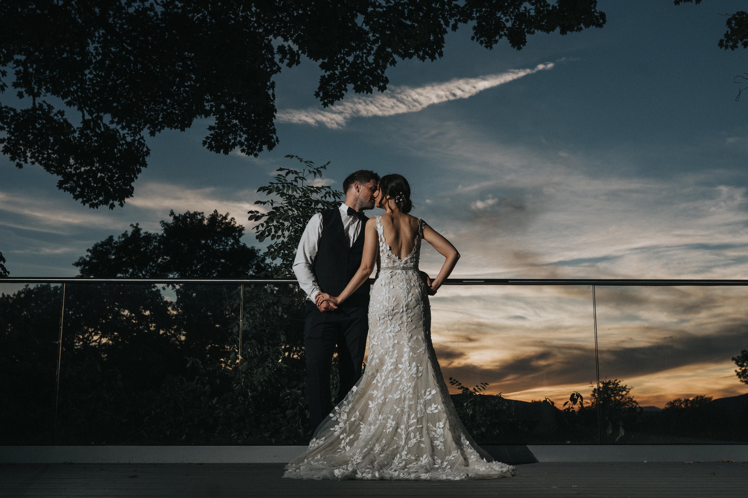 A bride and groom are standing close together outdoors at sunset, kissing beneath a sky with clouds and a sharp vapor trail, surrounded by trees.