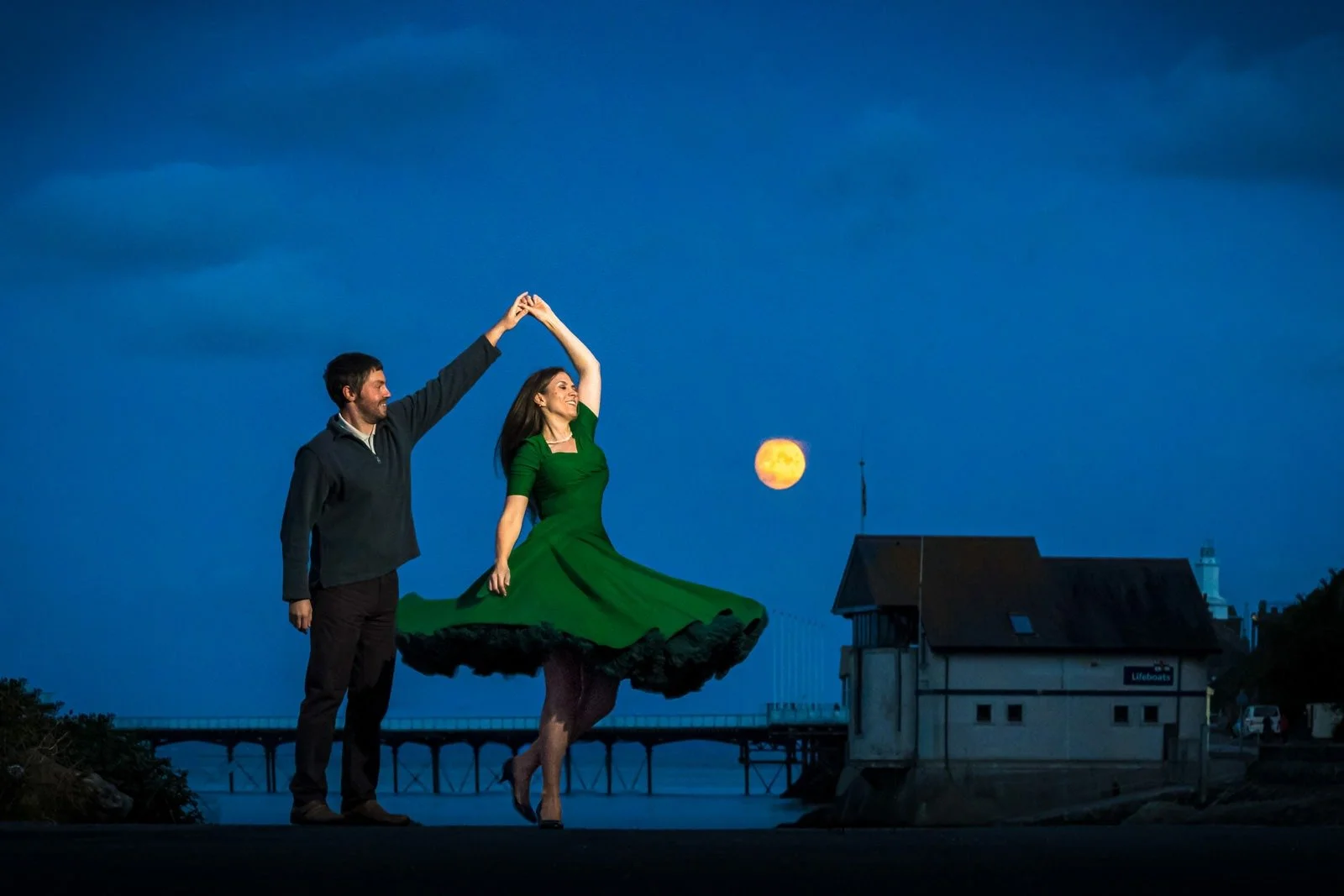 A couple dancing outdoors at twilight, with the woman in a green dress and the man in a dark shirt, holding hands above her head, and a bright full moon in the background next to a building by the water.