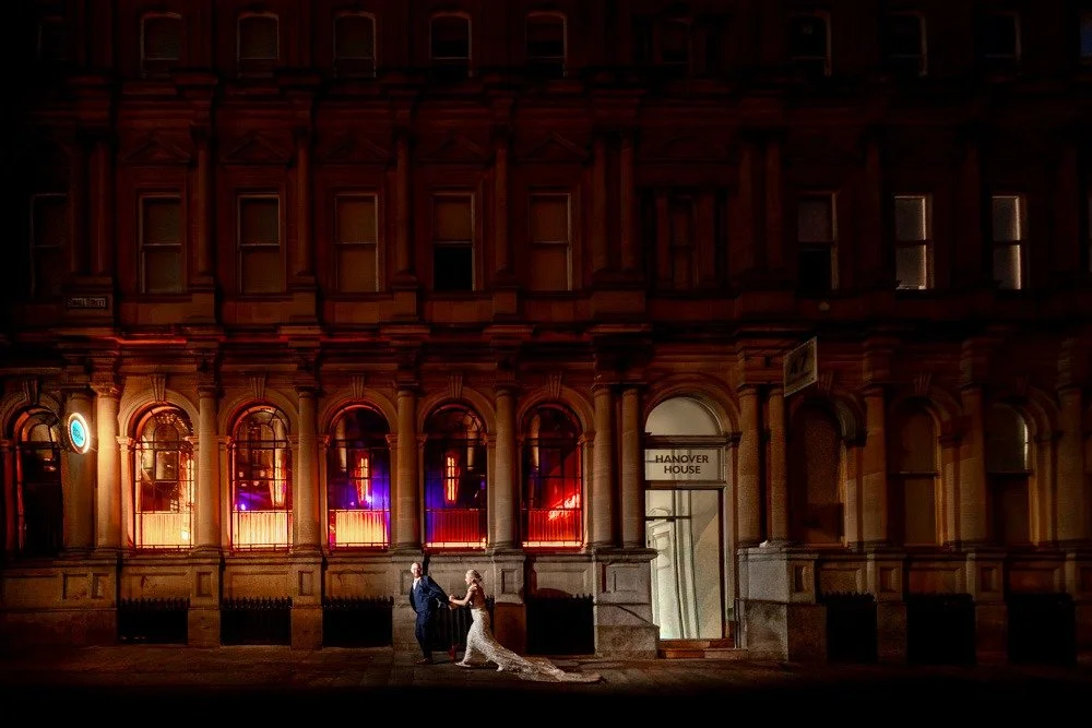 A couple in wedding attire holding hands, standing outside a building with the sign Hanover House, at night. The building has tall windows with colorful lights.