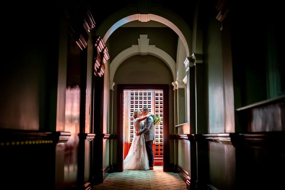 A bride and groom sharing a kiss in a vintage-style hallway with arched windows, dark wood paneling, and patterned carpet.