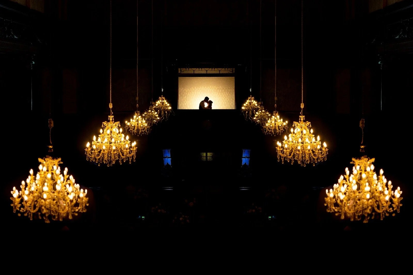 Silhouette of a couple kissing on a balcony with multiple ornate gold chandeliers hanging overhead in a darkened room.