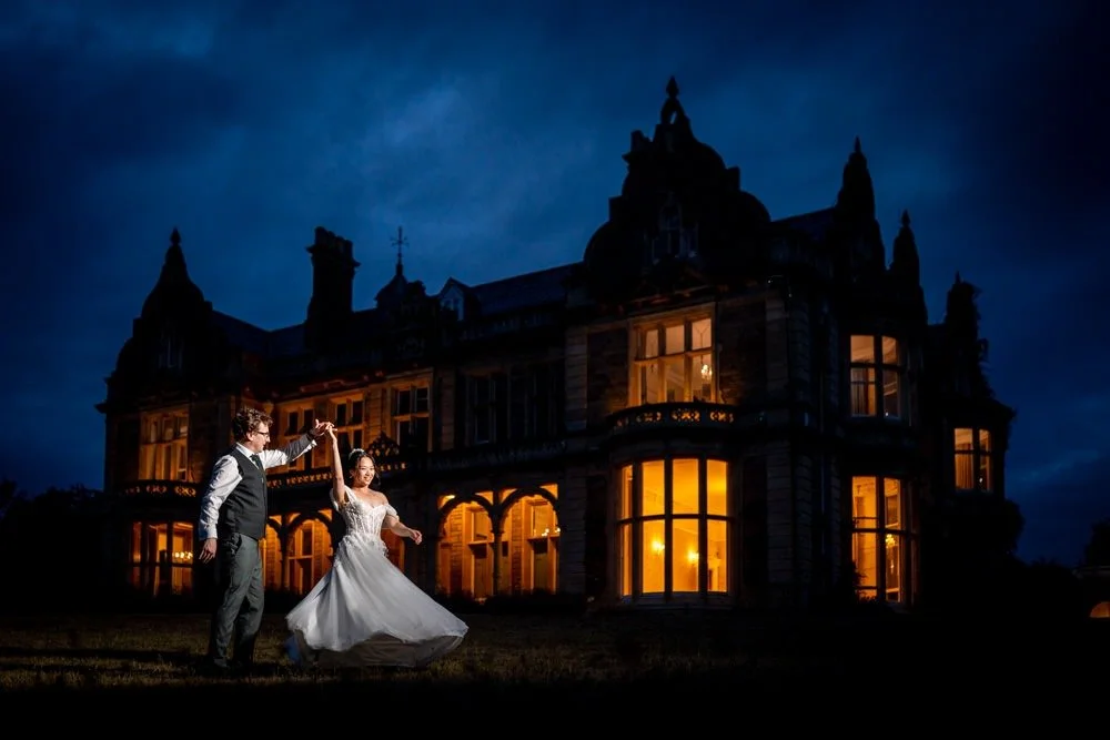 A newlywed couple dancing outside a large, illuminated mansion at night.