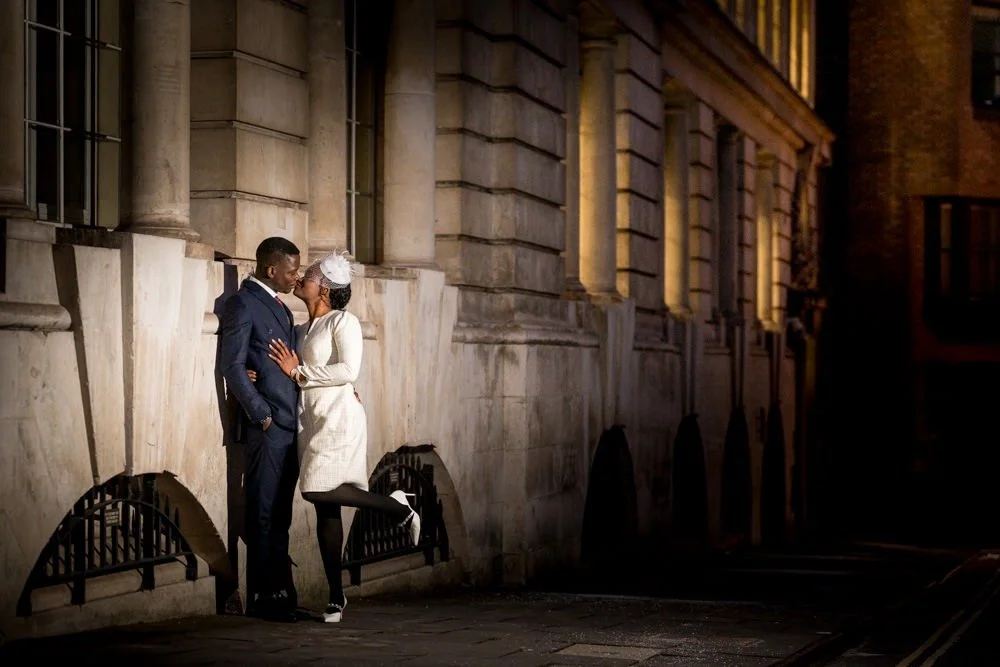 A couple dressed in formal attire sharing a kiss on a city street at night, standing against a stone building with warm lighting.