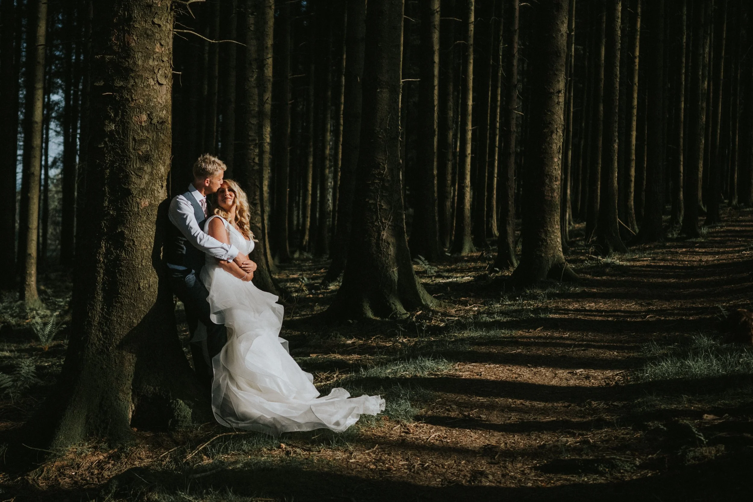 A bride and groom standing in a forest, embracing each other romantically. The groom is leaning against a tree while the bride stands close, looking up at him, with trees extending into the background.