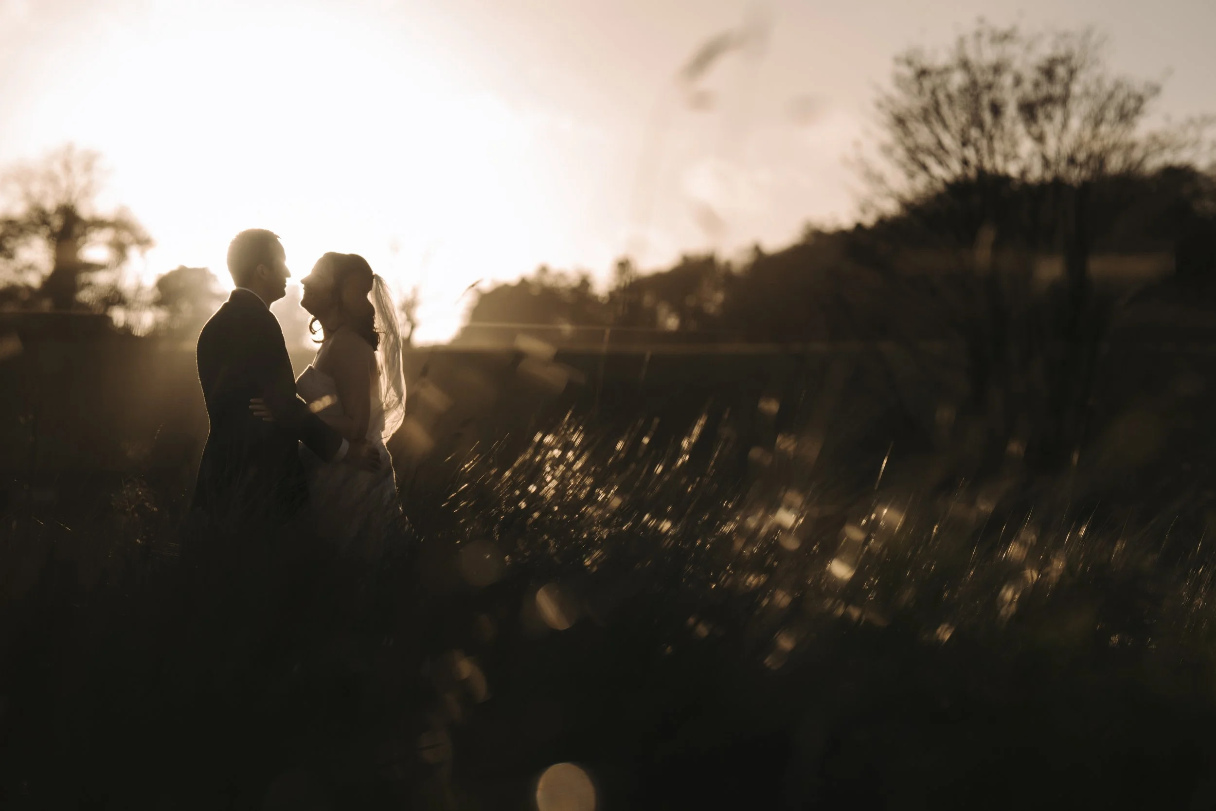 Silhouette of a bride and groom holding each other outdoors during sunset, with trees and a bright sky in the background.