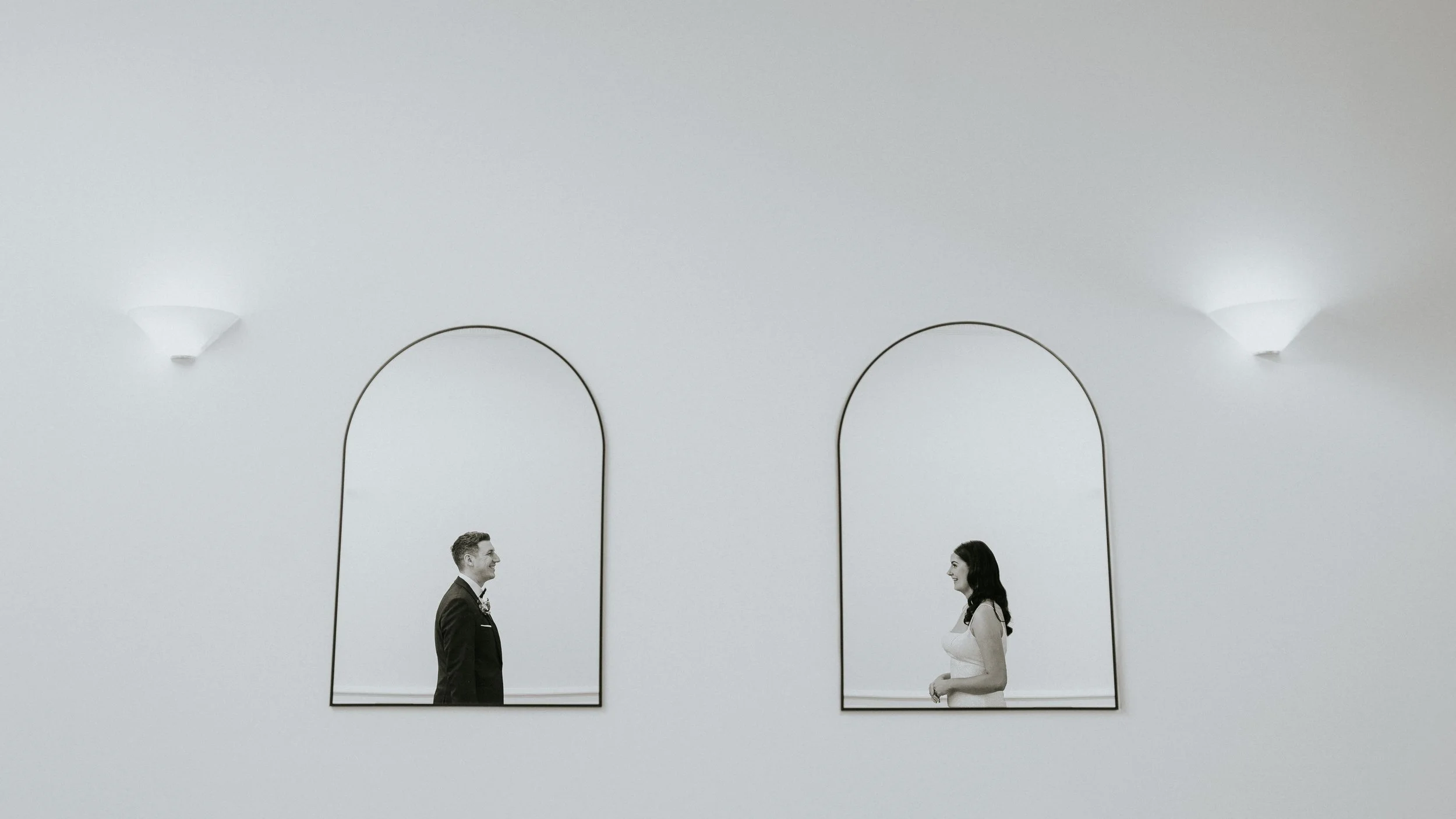 A man in a suit and a woman in a dress exchange smiles and look at each other in front of minimalist mirrors and wall sconces in a modern, white-walled room.