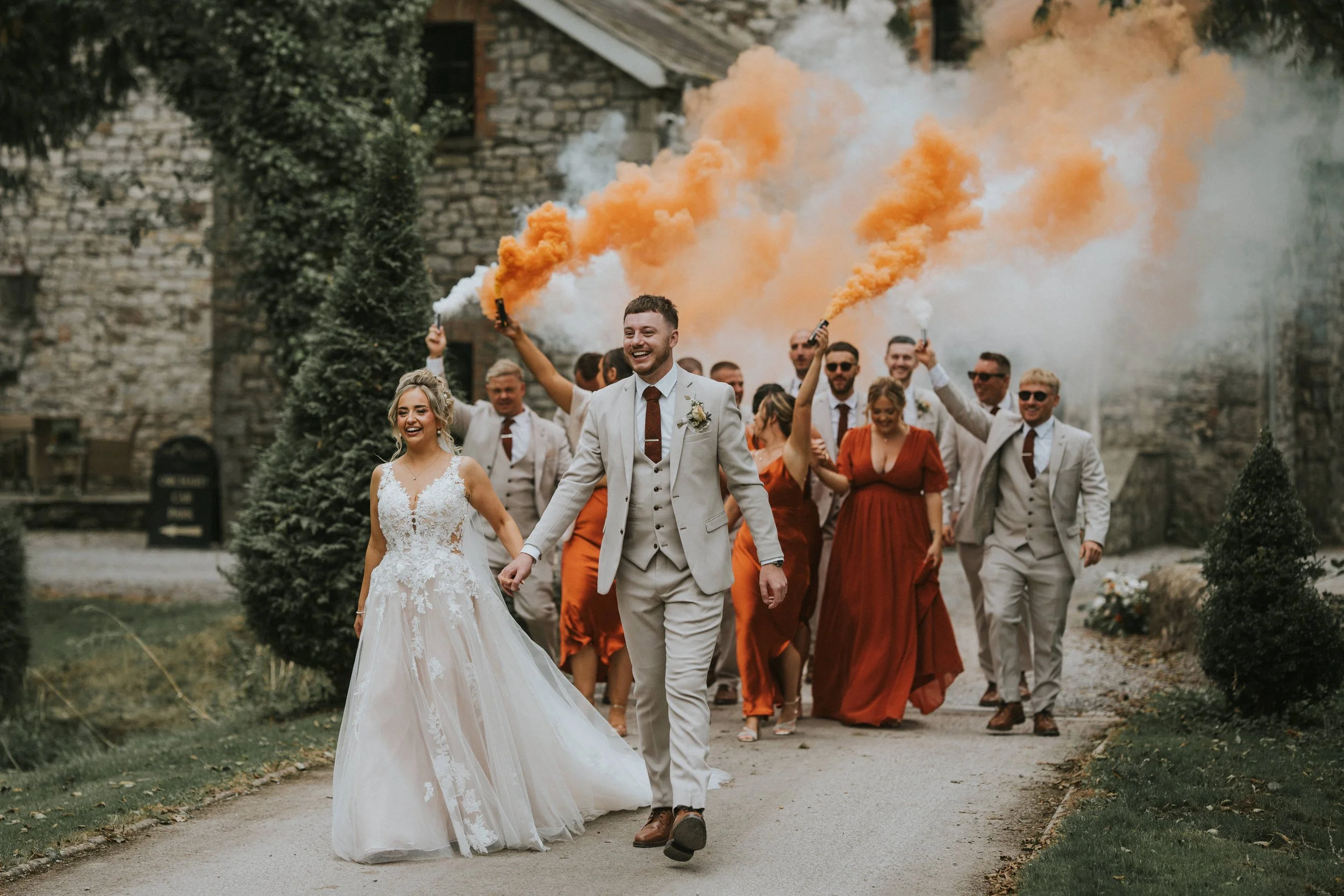 A wedding party walking outdoors, with the groom and bride at the front, smiling, as the guests behind them hold orange smoke grenades creating clouds of orange smoke.