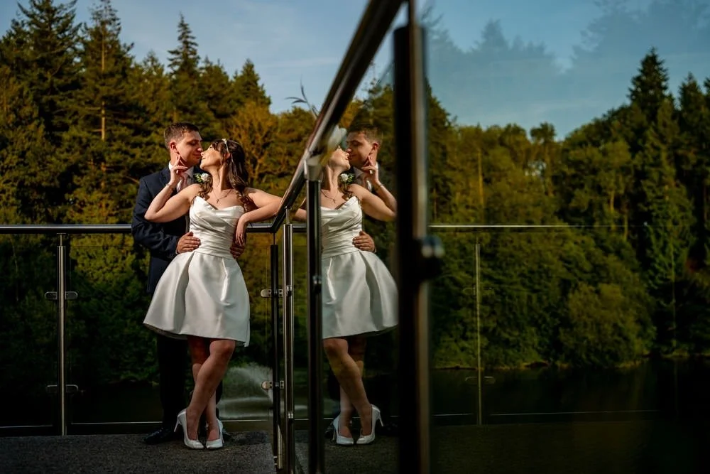 A couple dressed in formal attire standing on a balcony with a glass railing, surrounded by a forested area. They are sharing a kiss, with their reflection visible on the glass.