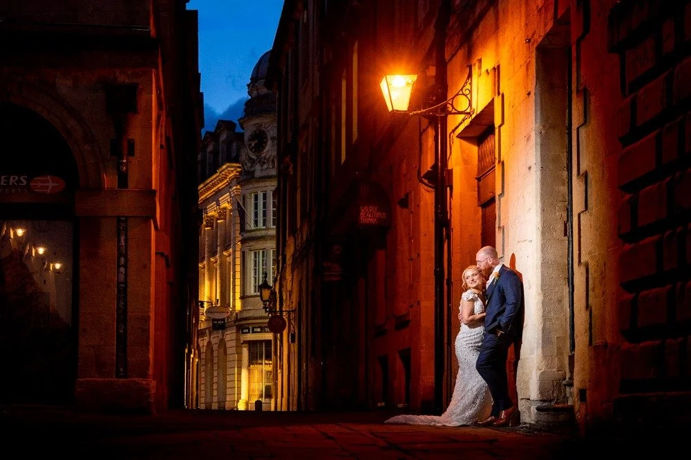 A couple standing close together in a narrow, dimly lit urban alley at night, with the woman in a wedding dress and the man in a dark suit, illuminated by a streetlamp.