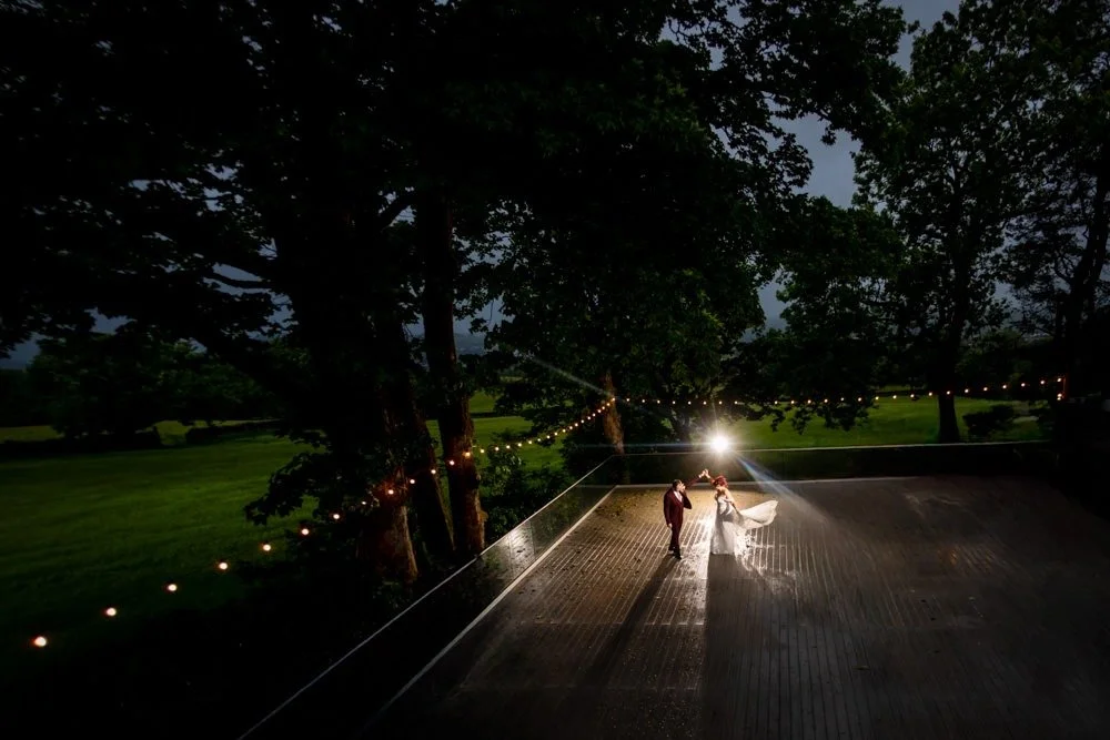 A bride and groom dancing outdoors at night under string lights, with a dark sky and green trees in the background.
