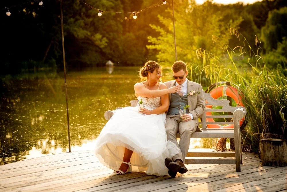 Couple in wedding attire sitting on a park bench by a lake with trees in the background during sunset.