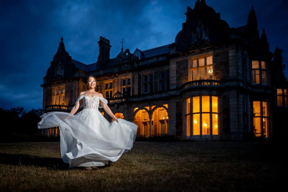 A woman in a wedding dress dancing outside a large illuminated castle-like mansion at dusk.