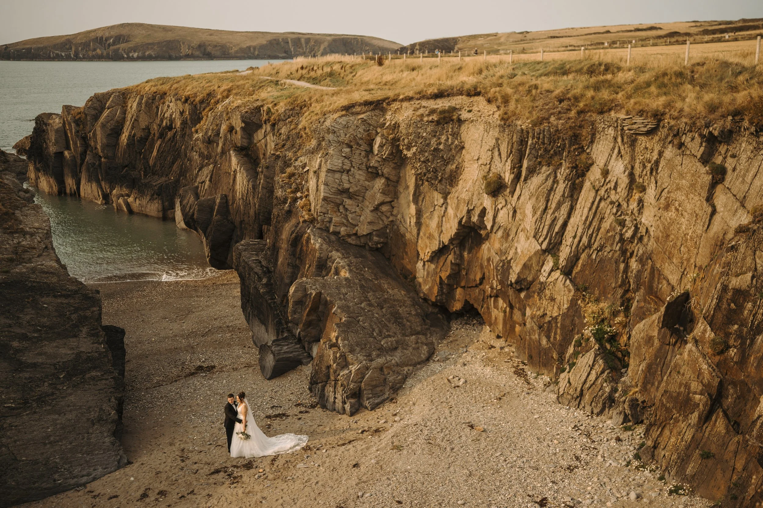 A bride and groom holding hands on a rocky beach under a cliff with grassy terrain and a fence in the background during sunset.