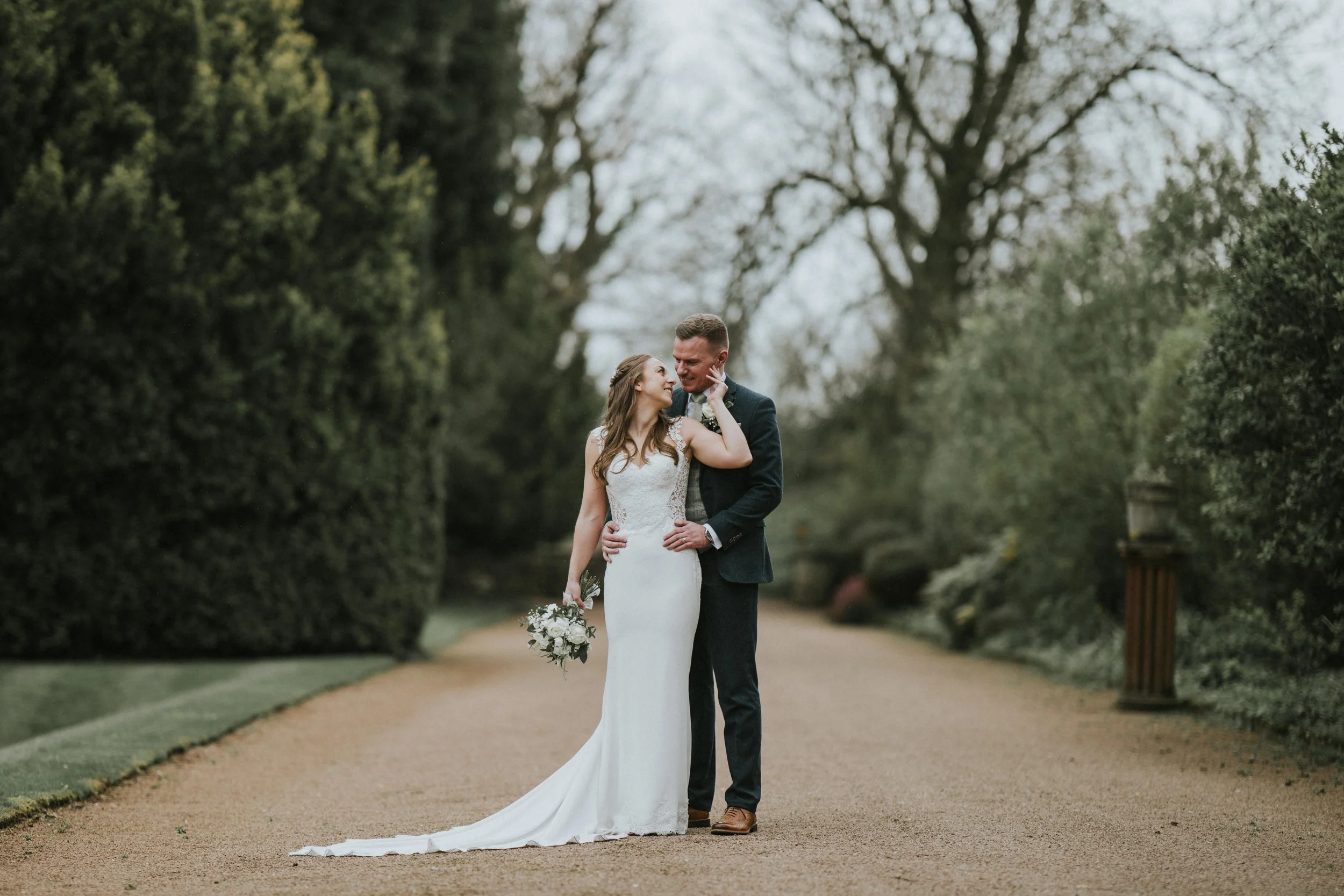 A couple in wedding attire walking outdoors on a path in a park-like setting, with the bride holding a bouquet of white flowers, and the groom in a dark suit.