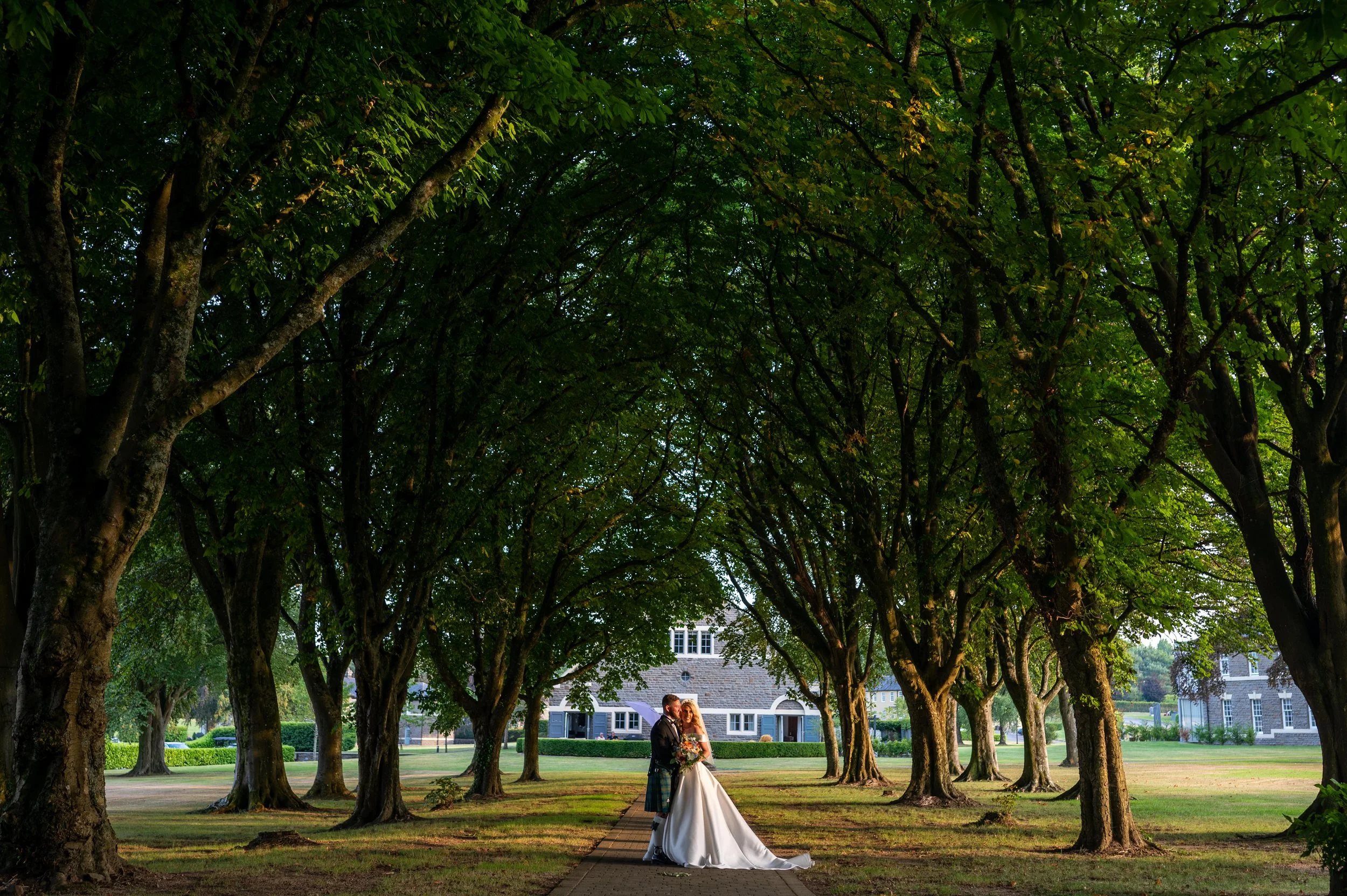 A bride and groom standing under a canopy of green trees during their wedding photoshoot, with houses visible in the background in a park-like setting.