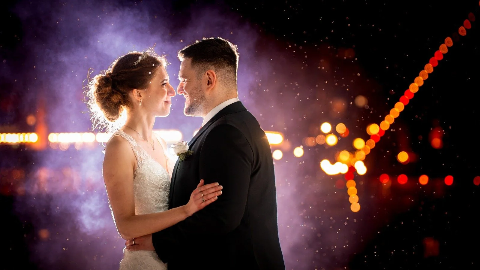 A bride and groom are close together, smiling and gazing into each other's eyes at night with blurred city lights and fireworks in the background.