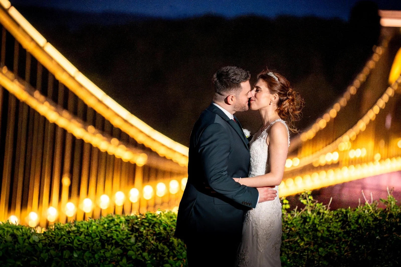 A newlywed couple kissing on a bridge at night, with lit strings of lights hanging across and greenery in the foreground.