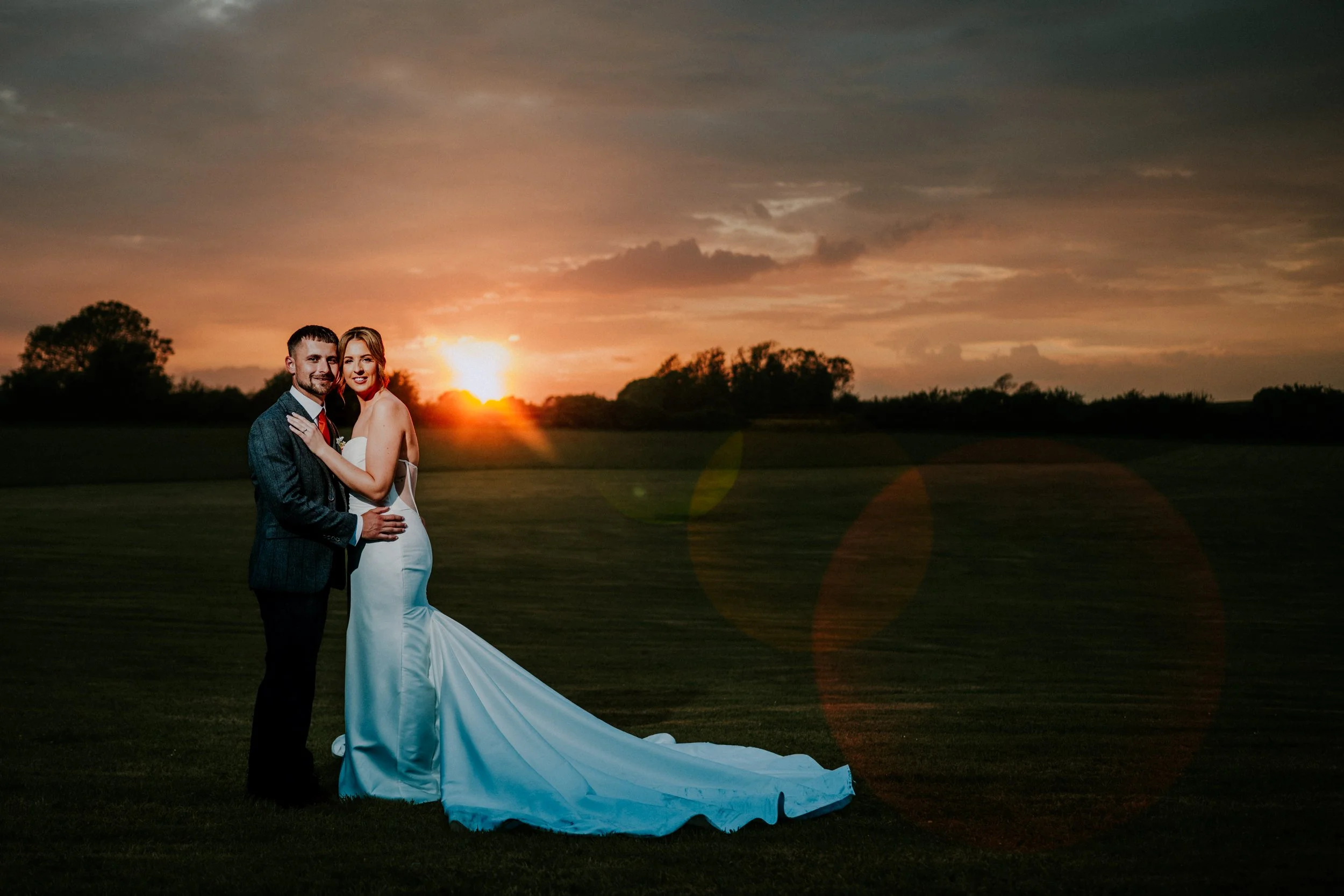 A bride and groom standing together outdoors during sunset. The bride is in a strapless white wedding gown with a long train, and the groom is in a dark gray suit with a white shirt and red tie. They are close, smiling, with the groom holding the bri