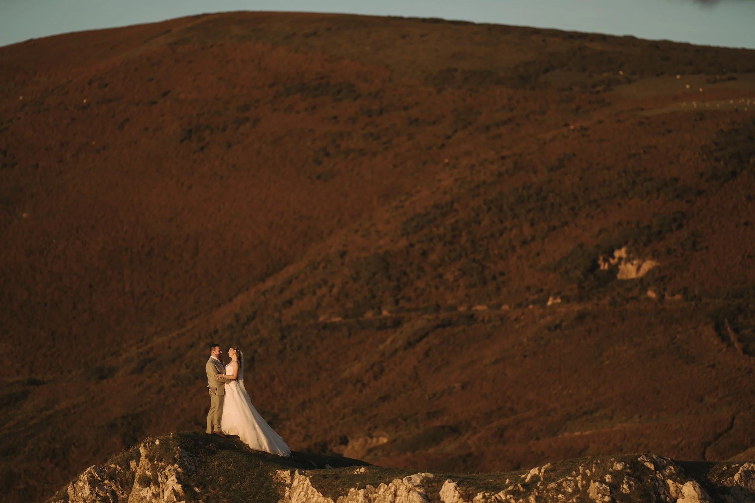 A bride and groom standing together outdoors on a rocky hill at sunset, with a large hill or mountain in the background.