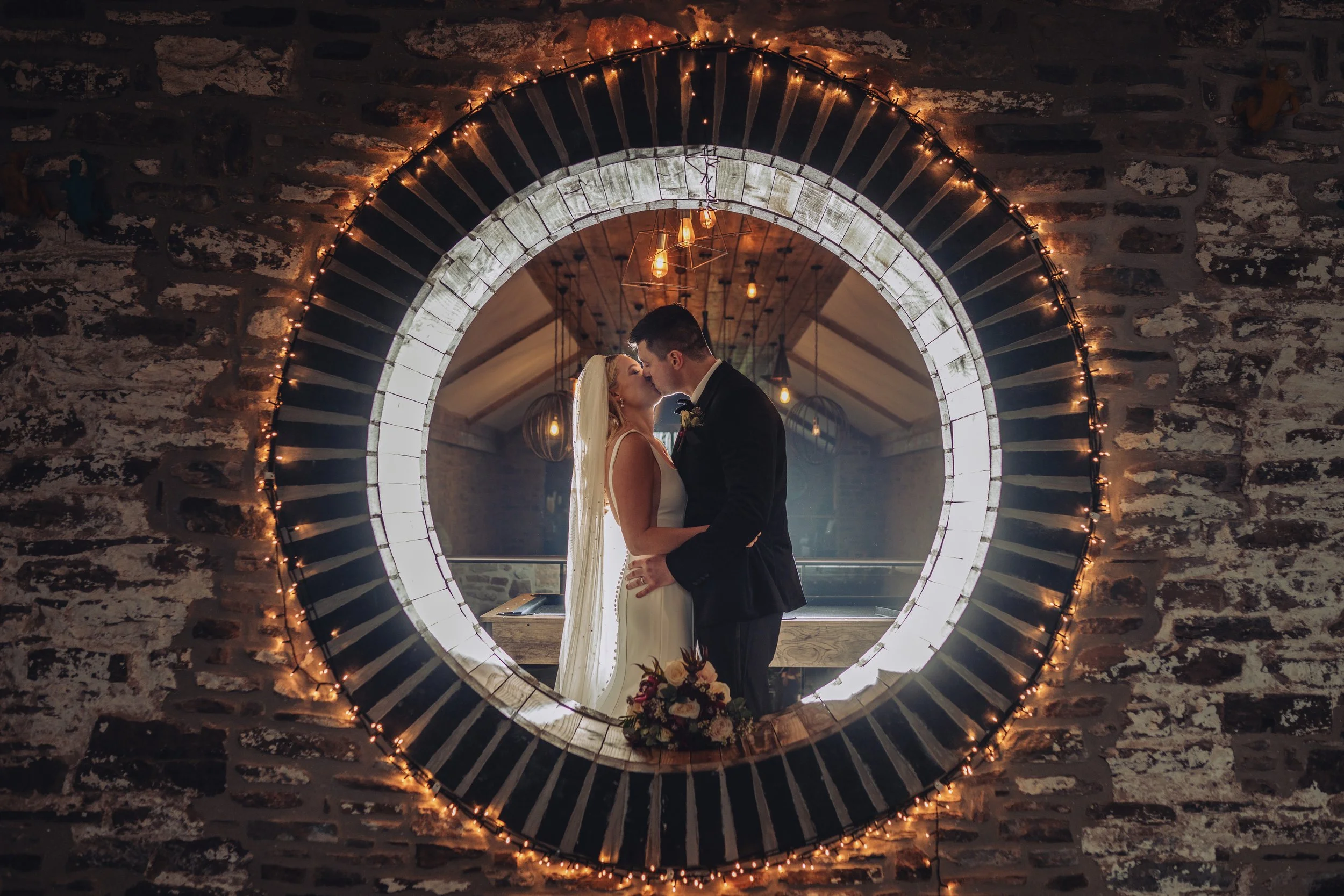 A couple in wedding attire sharing a kiss and holding each other inside a circular mirror framed with string lights, mounted on a brick wall.
