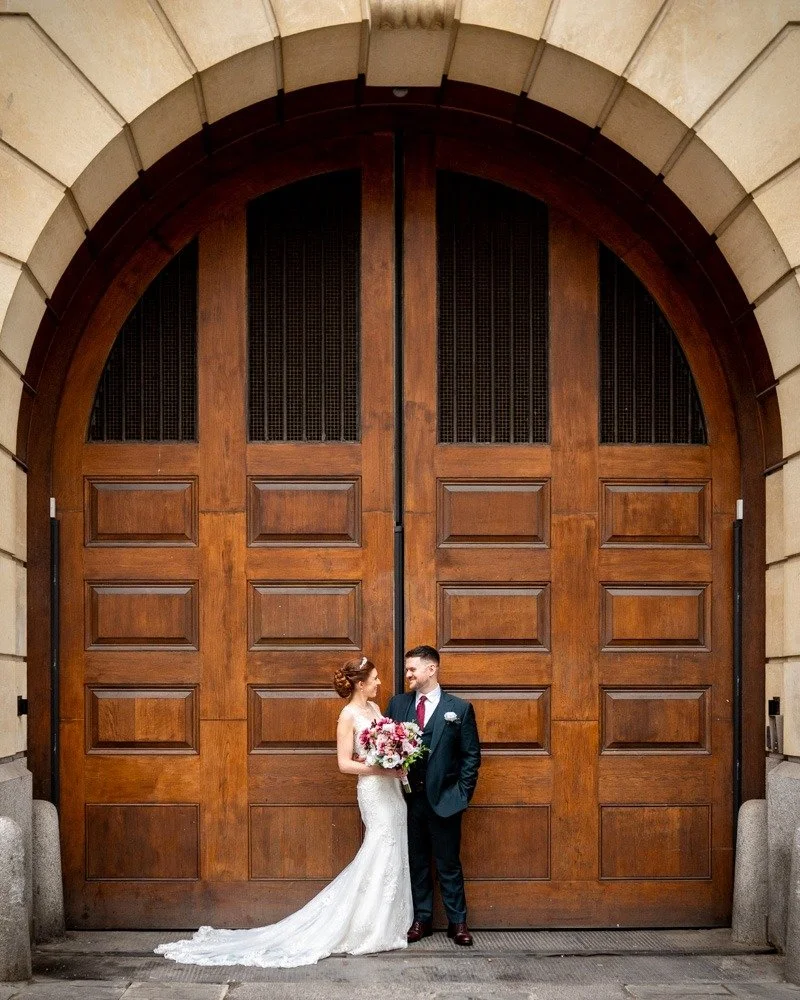 A bride in a white wedding gown holding a bouquet and a groom in a dark suit standing in front of large wooden double doors.