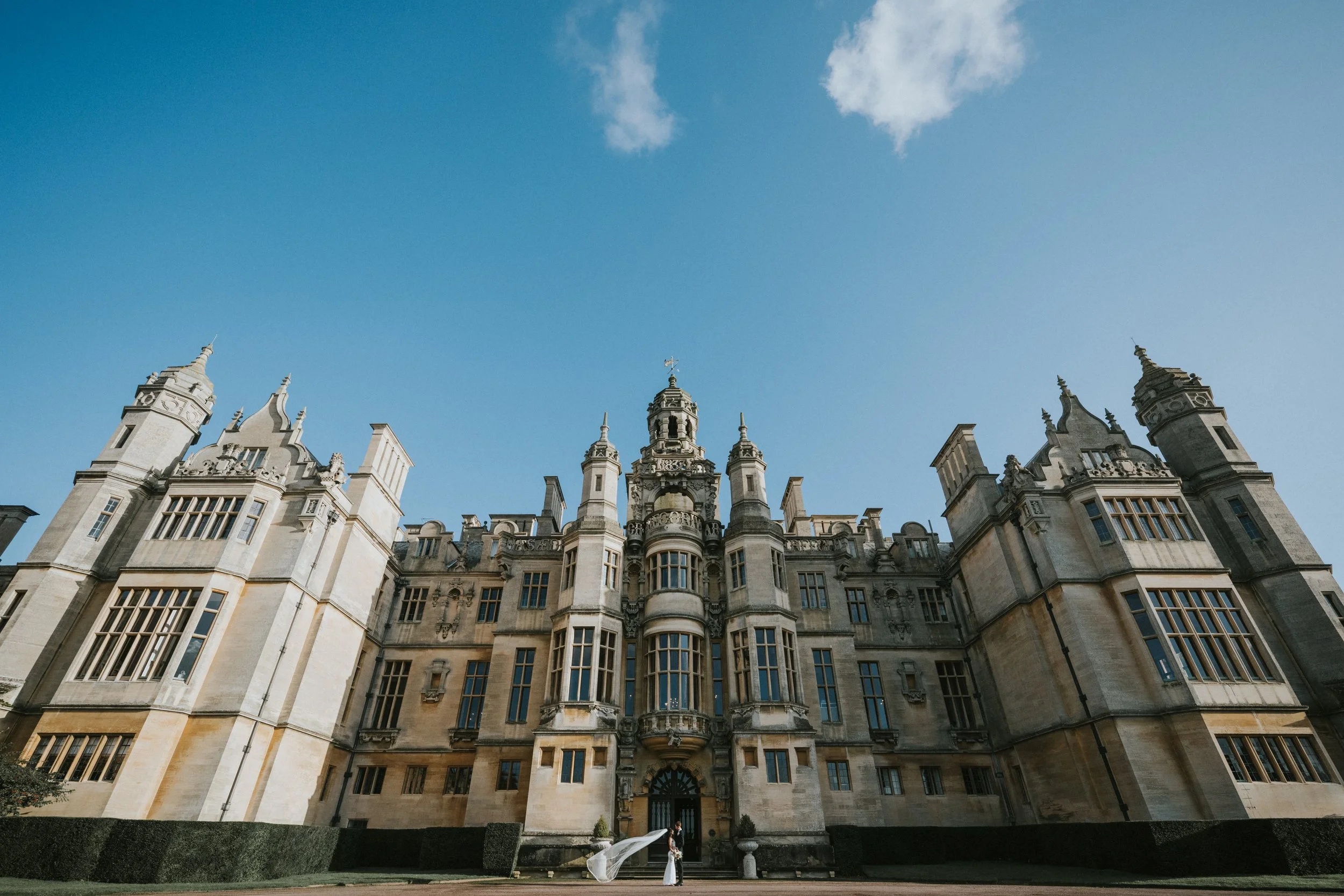 A large, historic stone building with ornate towers and turrets, set against a blue sky with some clouds.